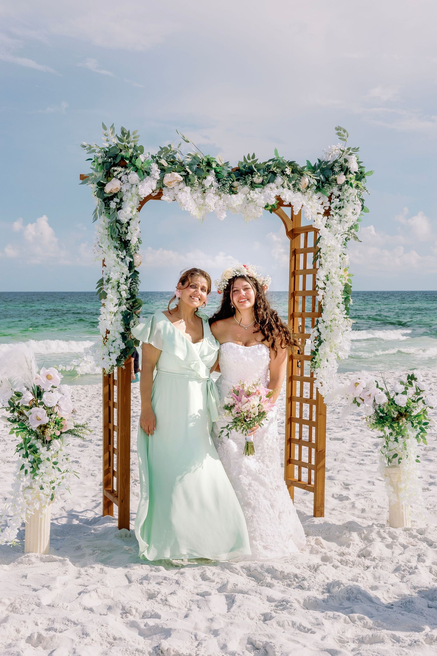 Two people in mint green and white gowns pose under floral beach wedding arch.