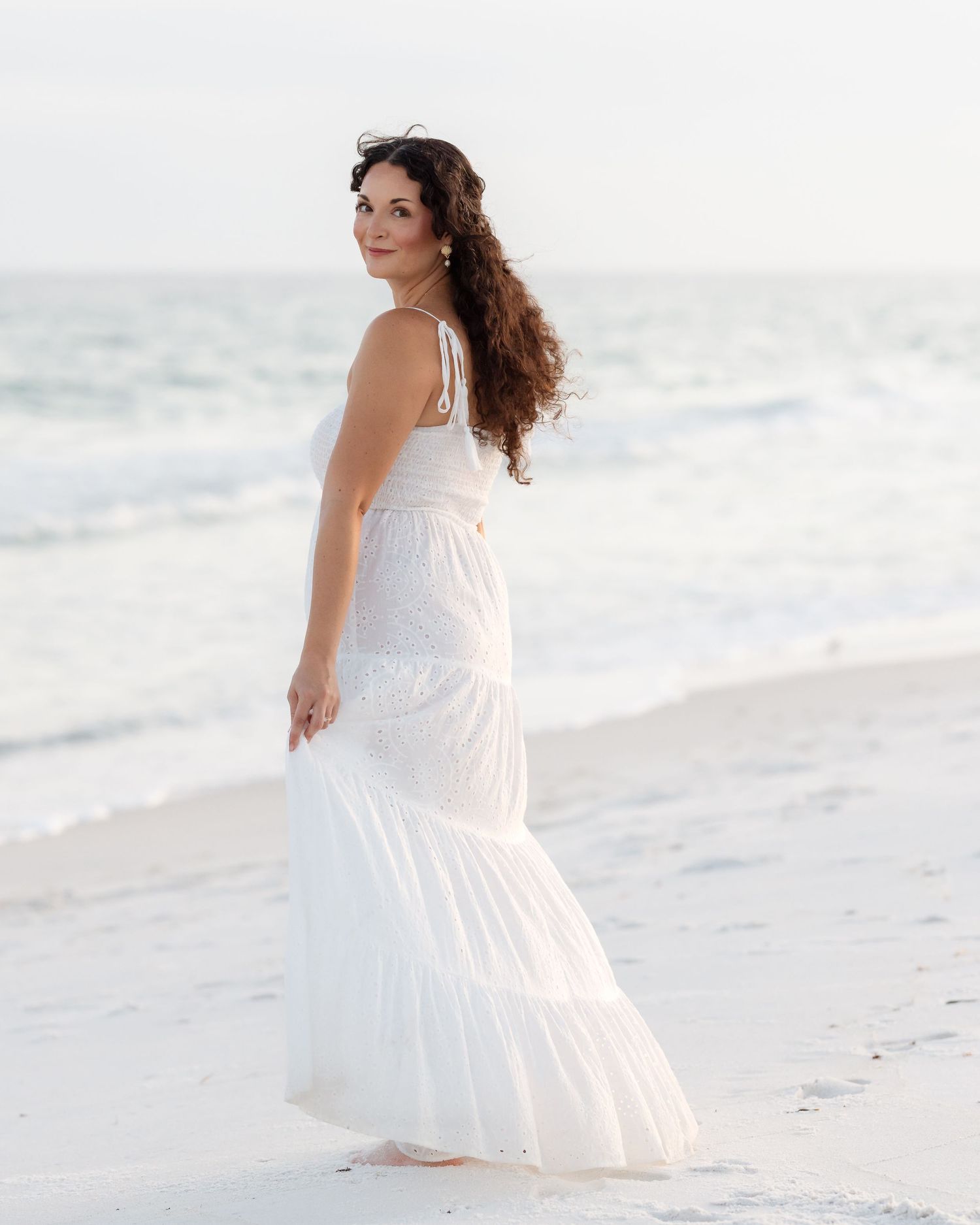 Woman in white maxi dress looking back over shoulder on beach.