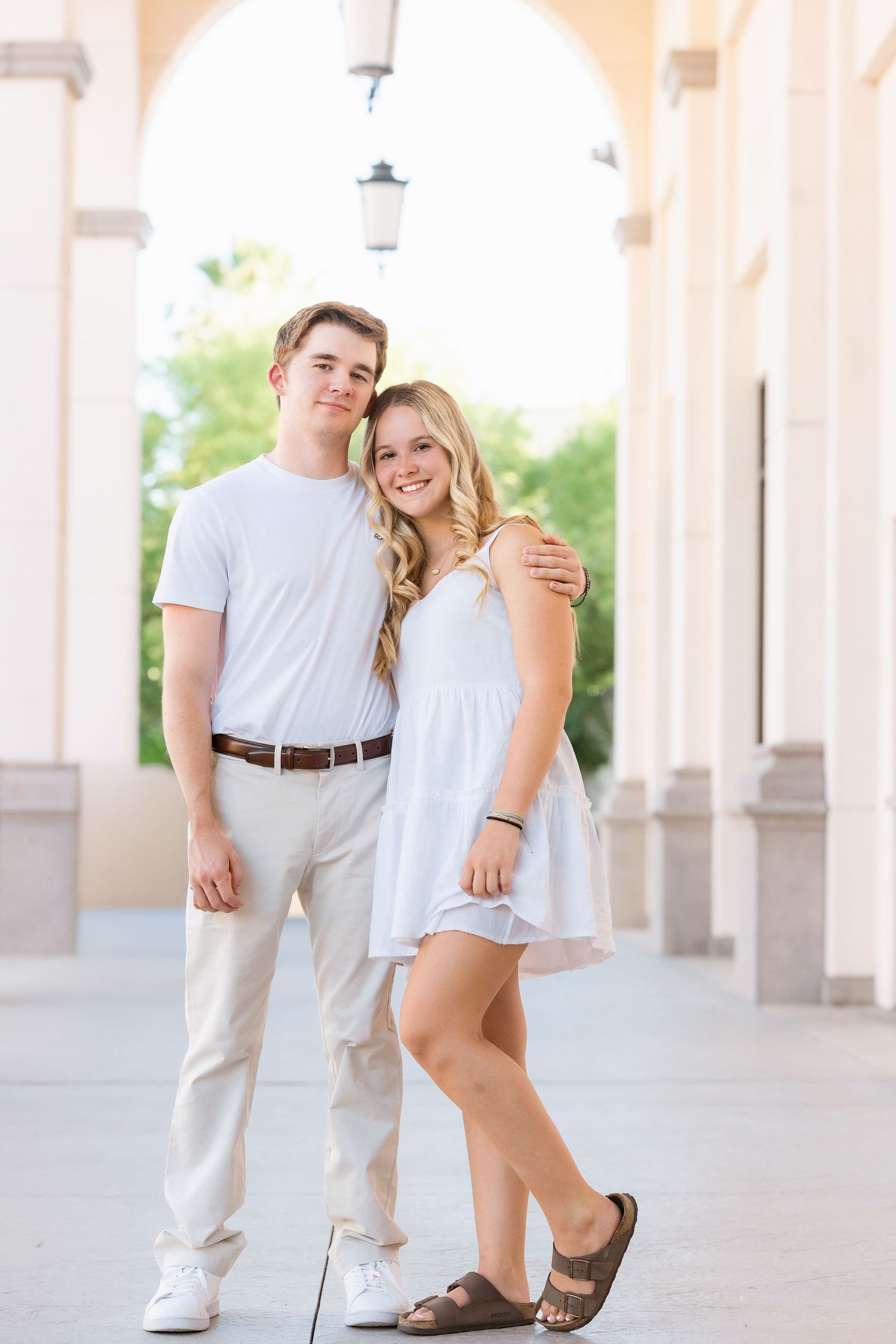 Young couple in white outfits poses together under arched columns in a bright outdoor setting.