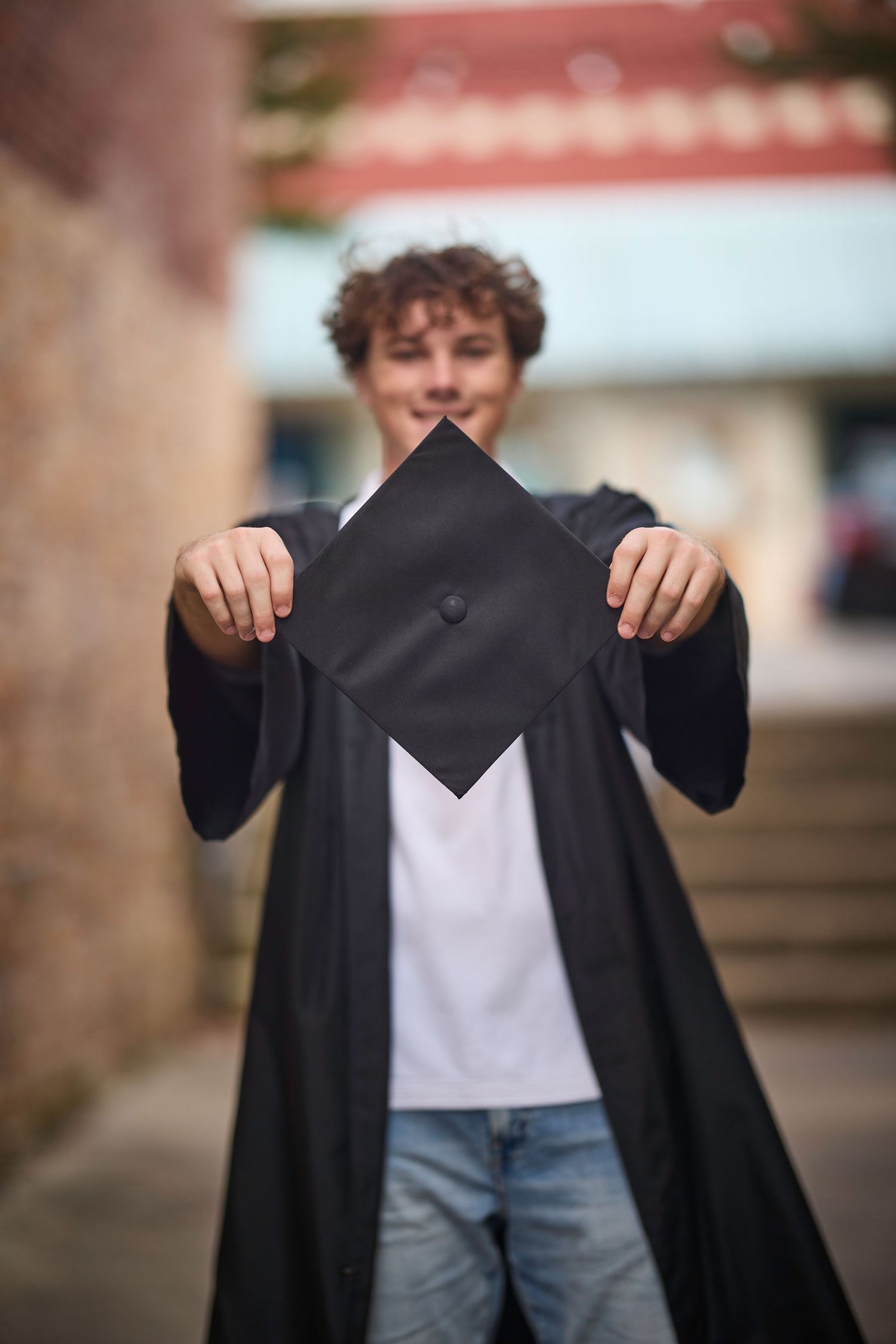 Young graduate in black gown holding mortarboard cap toward camera, smiling outdoors near brick building.