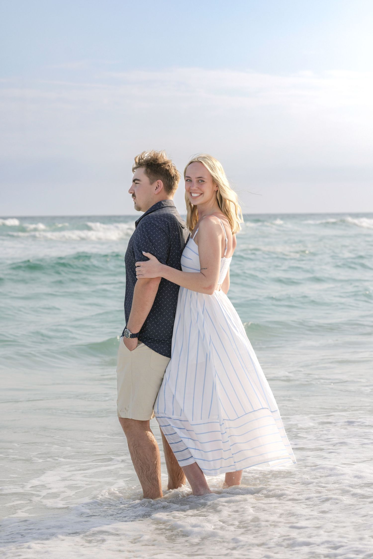 Happy couple embracing in ocean waves, woman in striped dress smiling at camera on sunny beach.