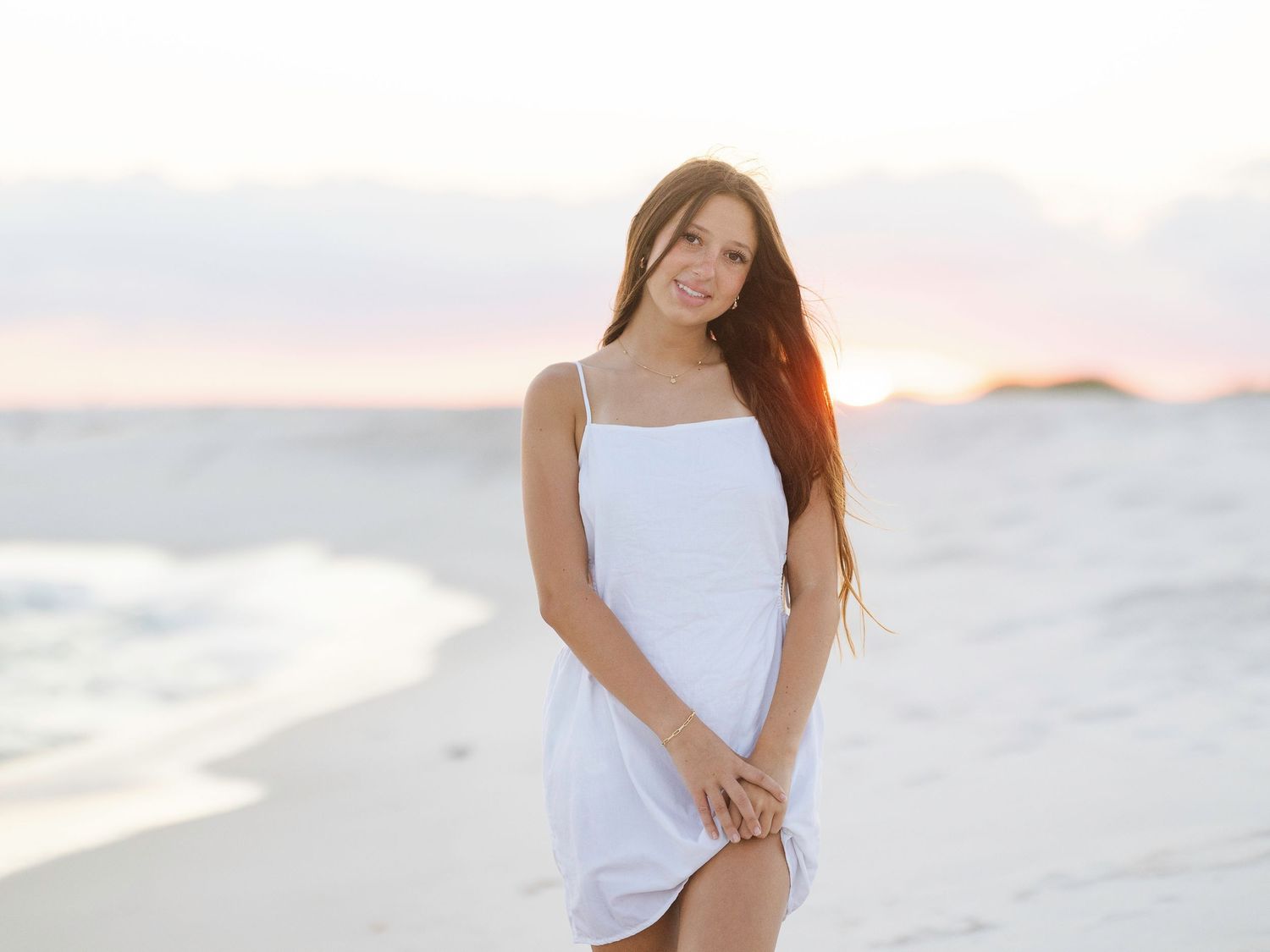 A person in a white sundress poses on a sandy beach at sunset with soft pastel colors in the background.