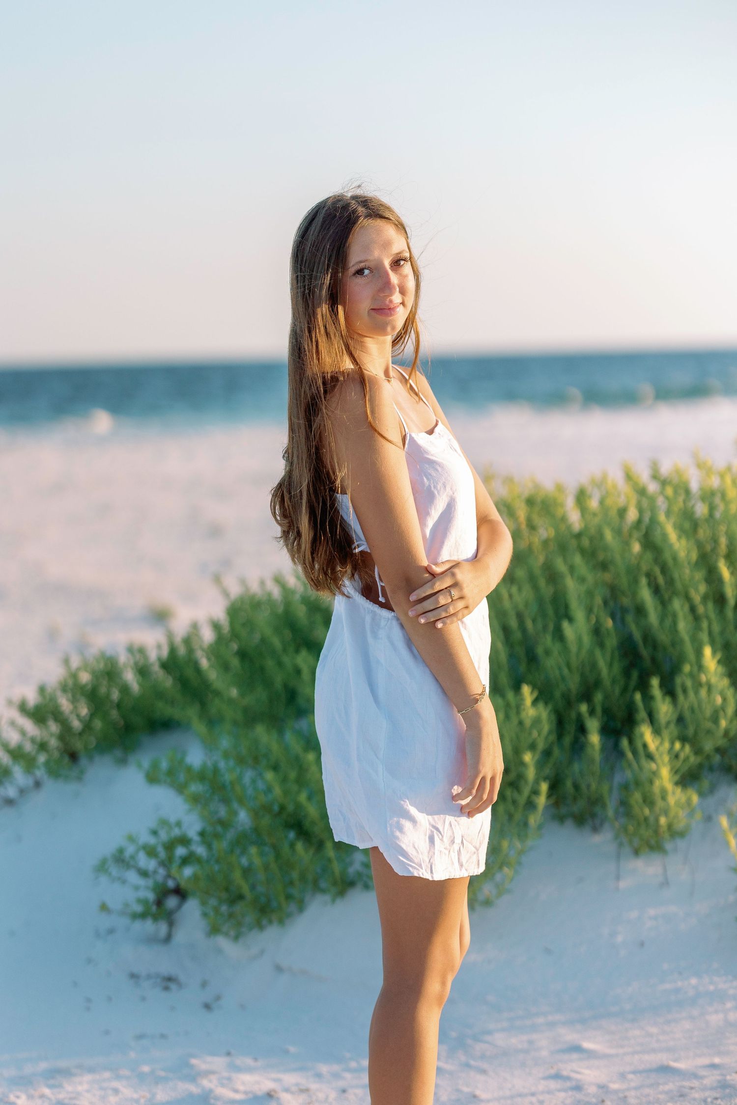 Woman in white outfit enjoying a peaceful moment on sandy beach with ocean backdrop.