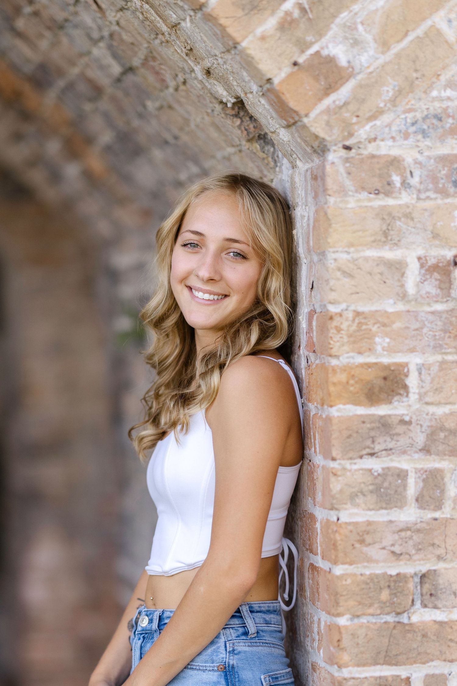 A woman in a white tank top and light jeans poses against a rustic brick wall in natural lighting.