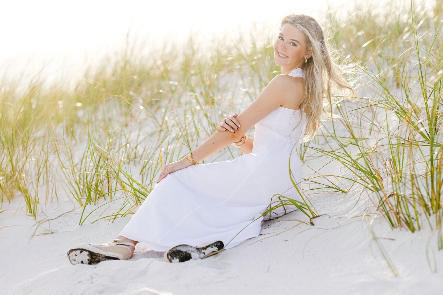A person in a white dress poses gracefully on a sandy beach with tall grass in the background.
