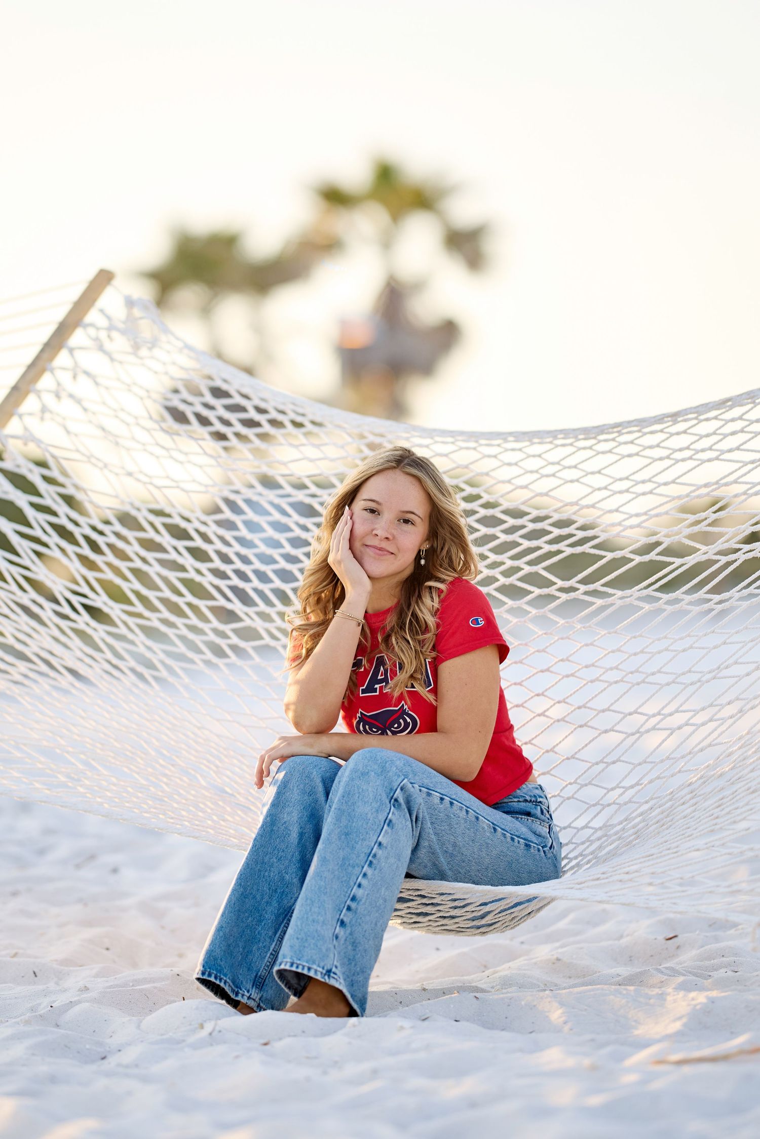 Person in jeans and red top relaxes in a white hammock on a sunny beach with palm trees in background.
