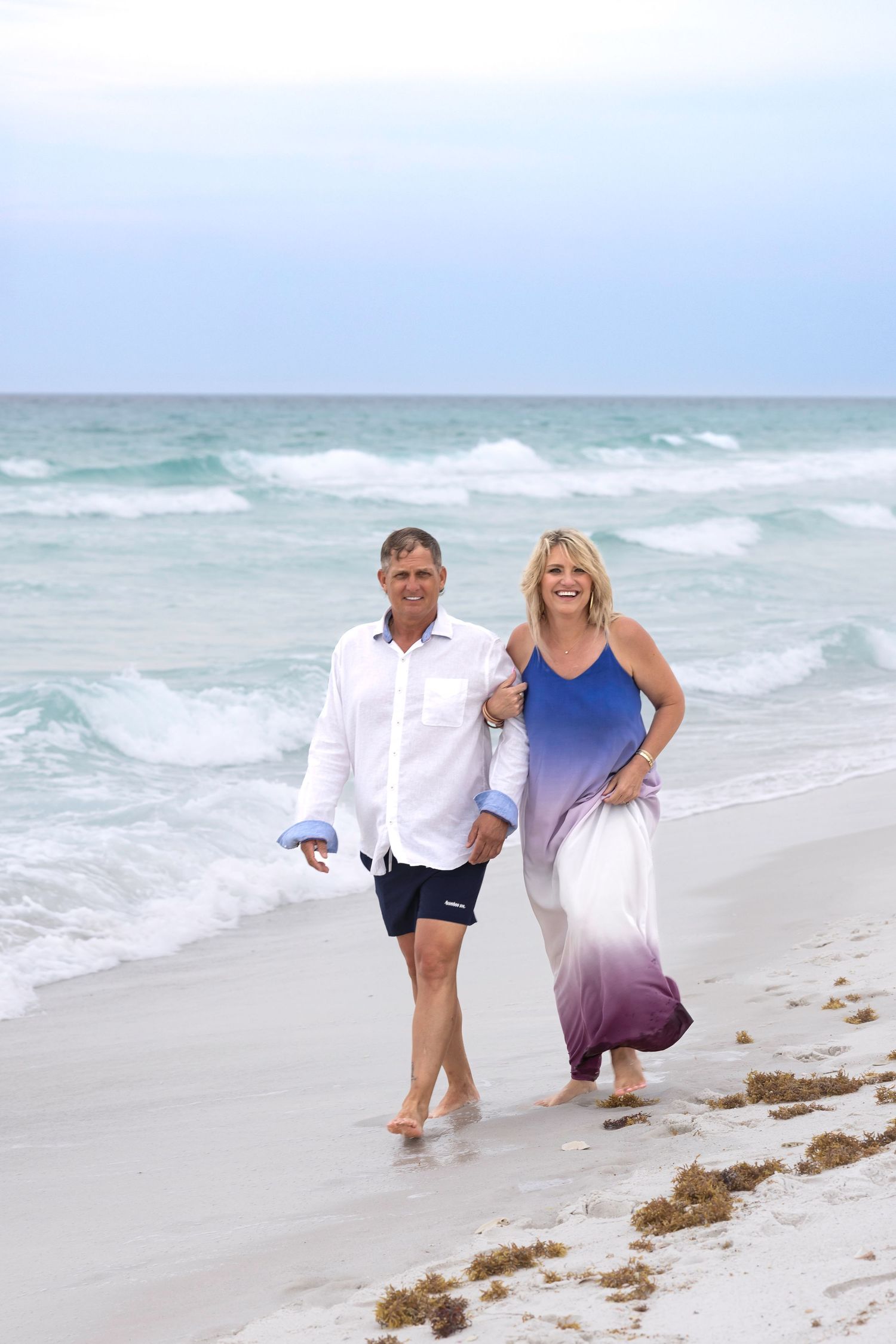 Happy couple walking hand in hand on a sandy beach with turquoise ocean waves in the background.