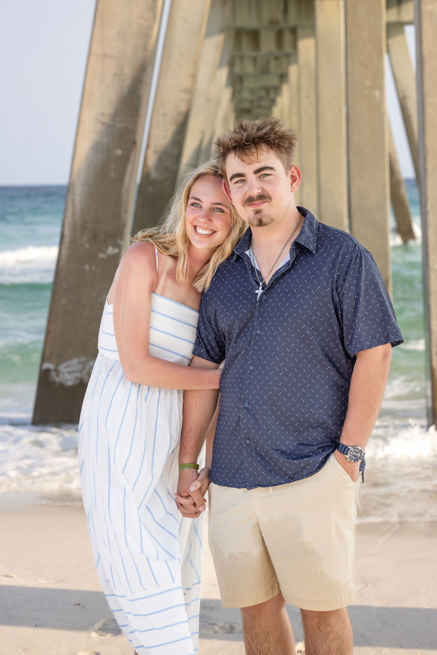 A smiling couple poses hand-in-hand on a beach near a pier, wearing summer outfits with ocean waves behind them.