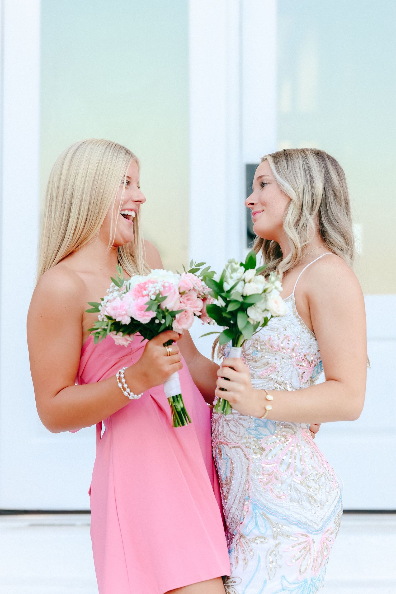 Two bridesmaids in pink and floral dresses share a joyful moment with bouquets at a wedding venue.