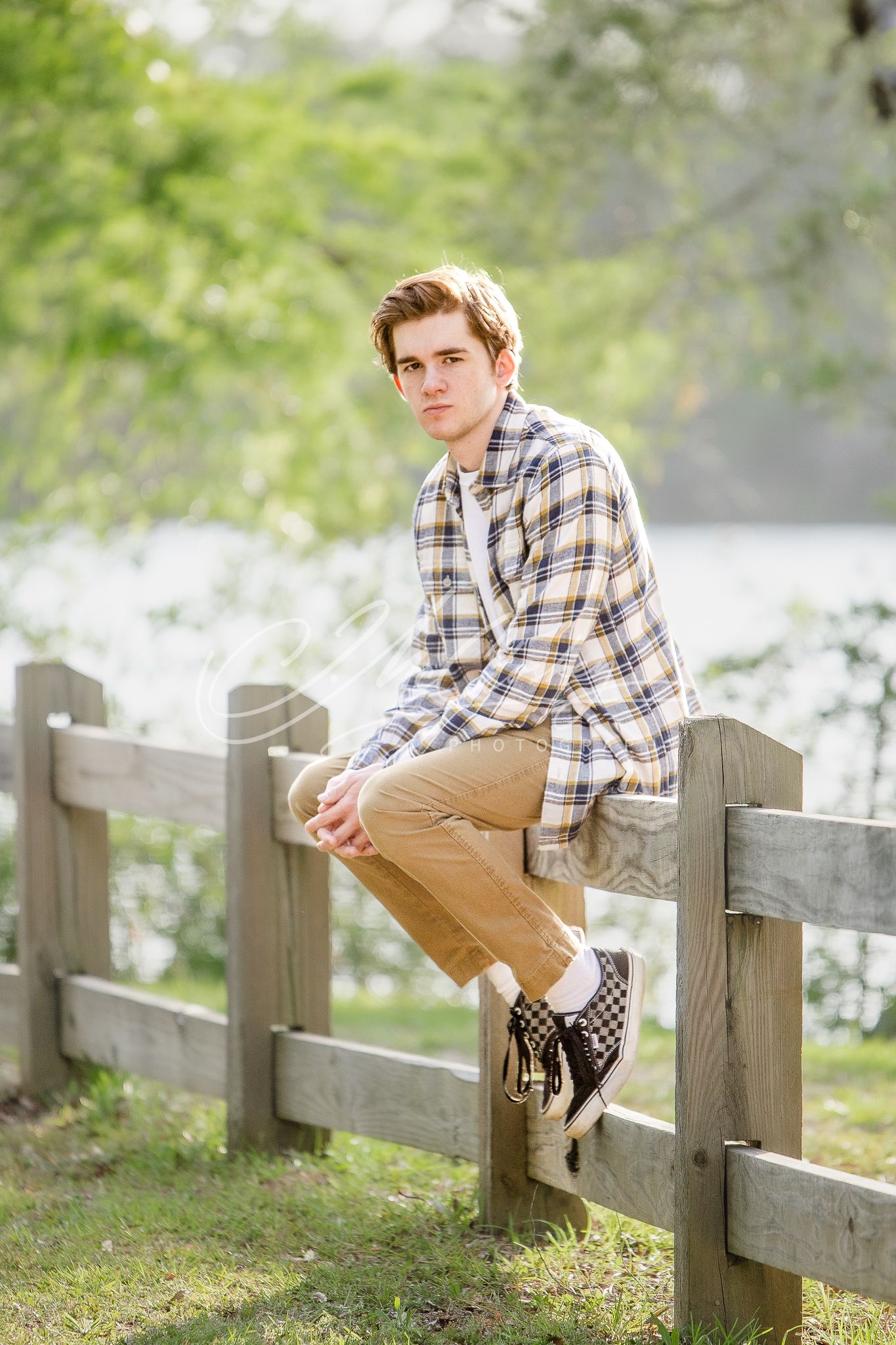A person in plaid shirt and khaki pants sits casually on a rustic wooden fence in a sunlit outdoor setting.