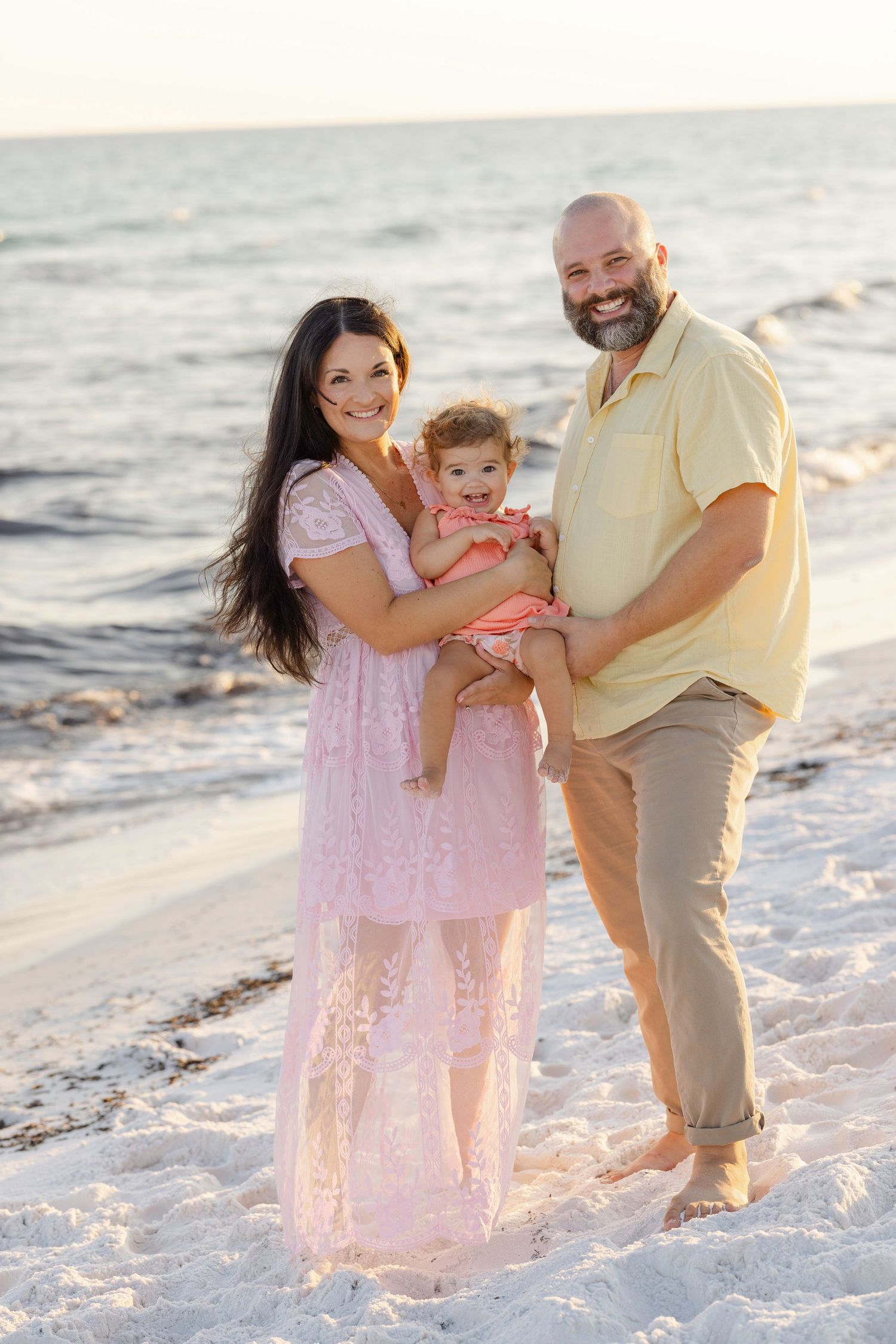A family poses together on a white sandy beach at sunset wearing coordinating pink and yellow outfits.