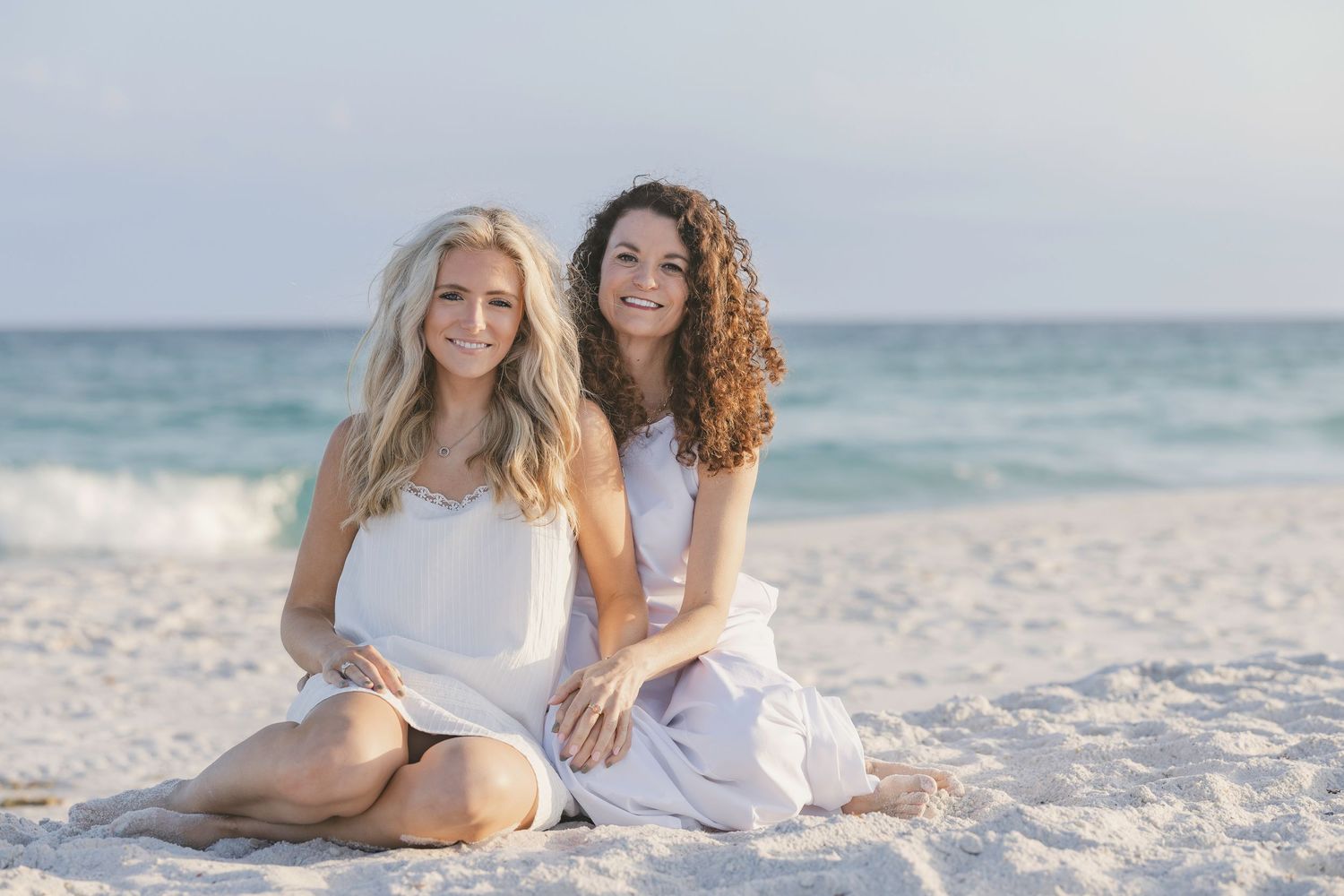 Two smiling women in white outfits sitting together on a sandy beach with ocean waves in the background.
