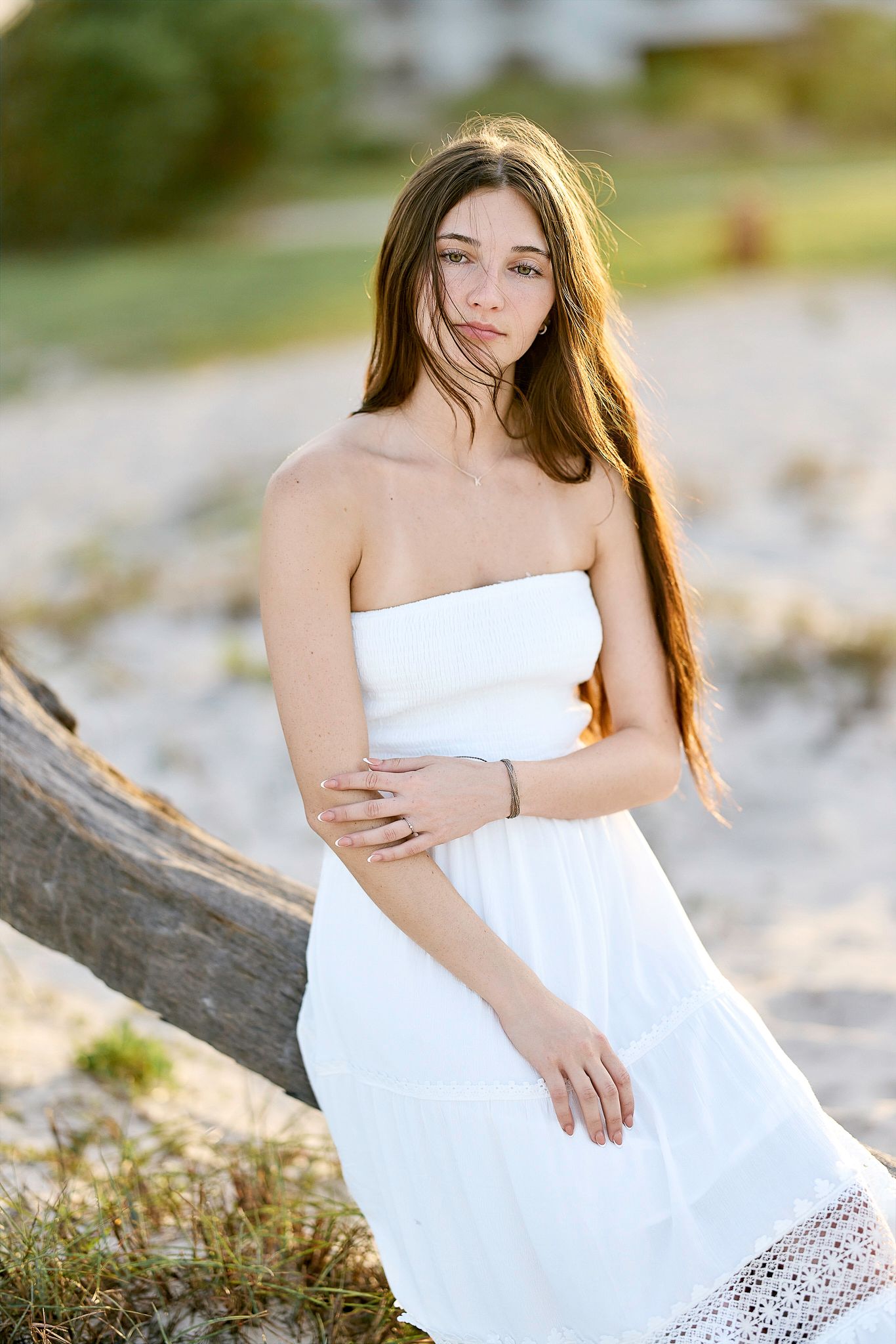 Young woman in white strapless dress posing by a palm tree on a sandy beach at golden hour.