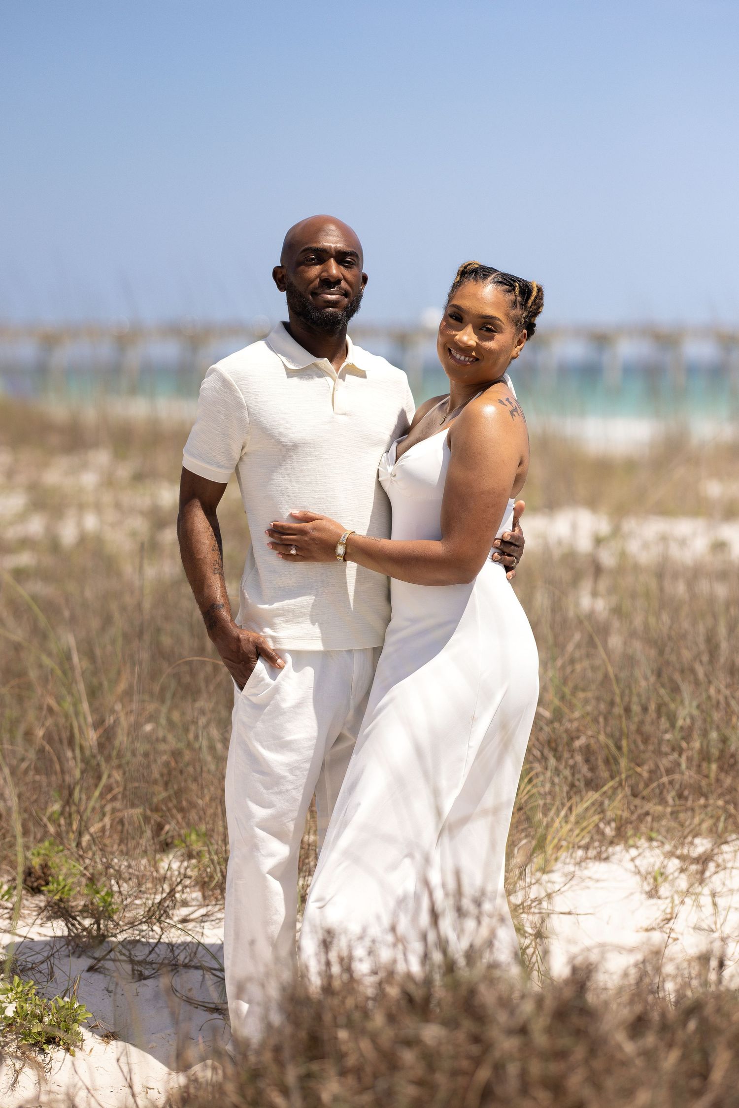 A couple in matching white outfits poses together on sandy beach dunes with ocean in the background.