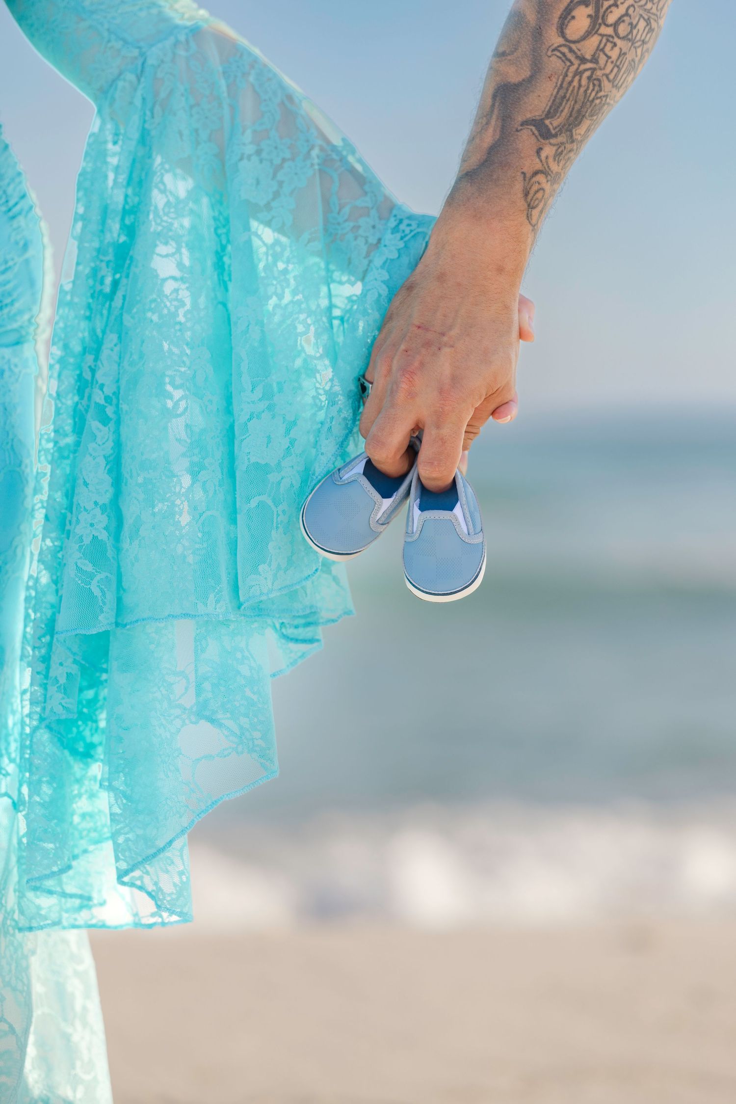 Tattooed hand holding tiny blue baby shoes near a teal lace dress on a sunny beach.
