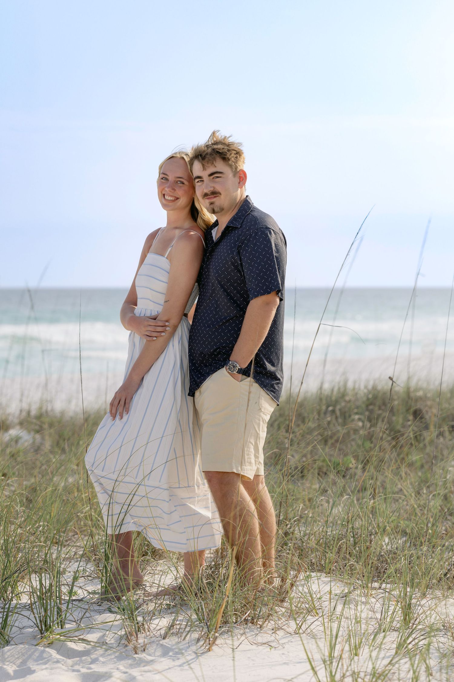 A young couple embracing on a sandy beach with sea grass, ocean waves, and blue sky in the background.