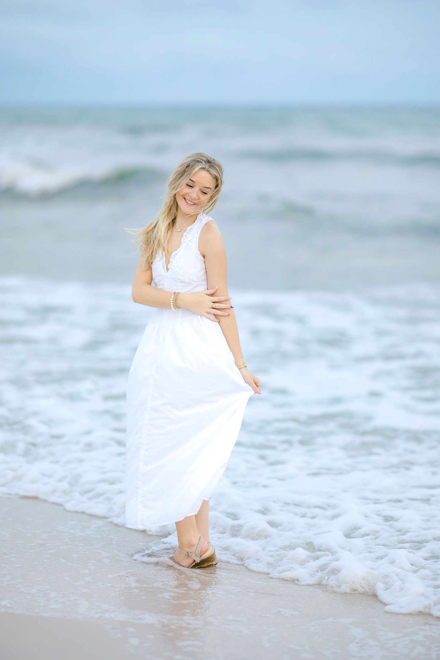 A smiling blonde woman in a flowing white dress stands at the ocean's edge, waves washing over her feet on a sandy beach.