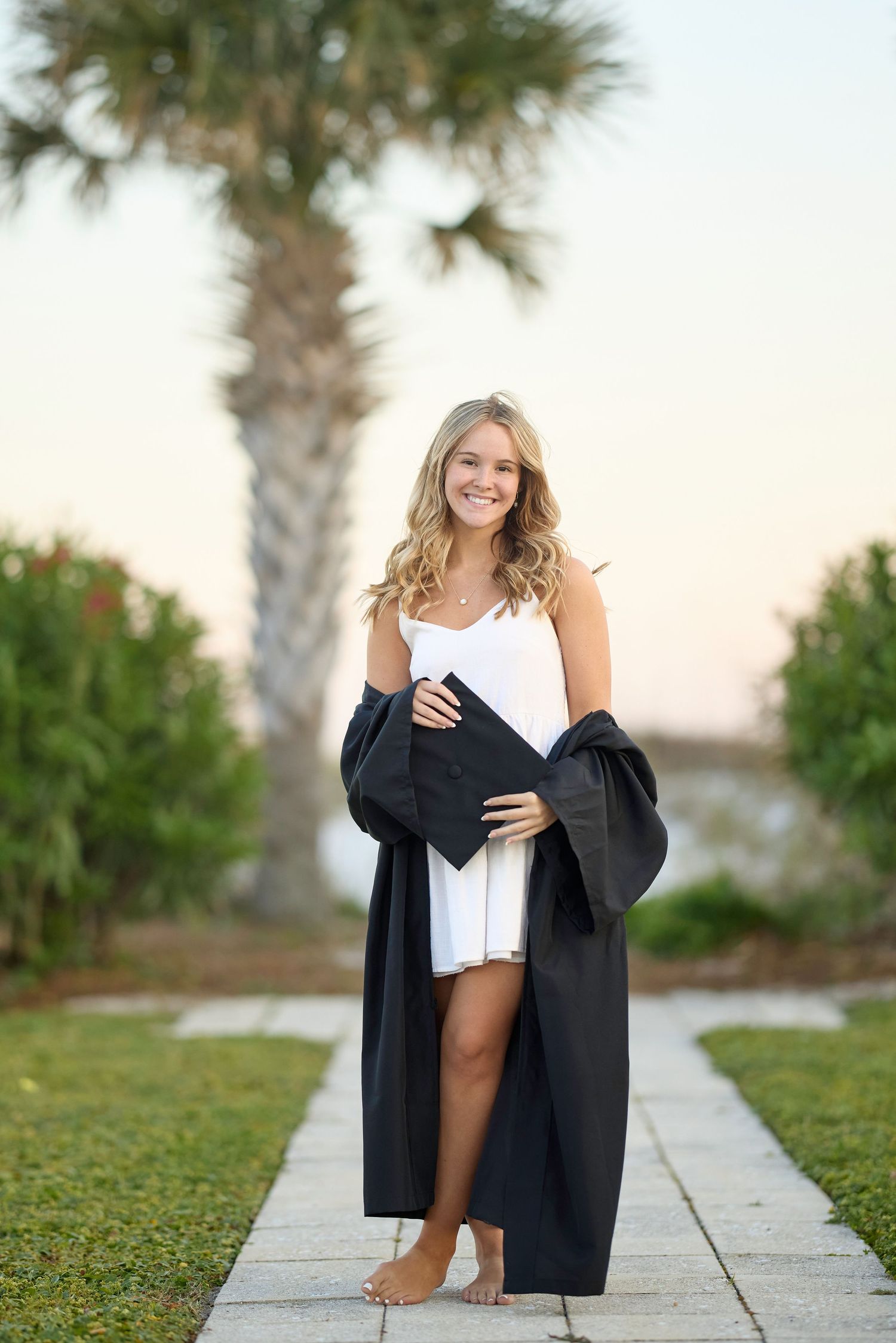Smiling blonde graduate in white dress and black cap and gown poses on garden path with palm tree backdrop.
