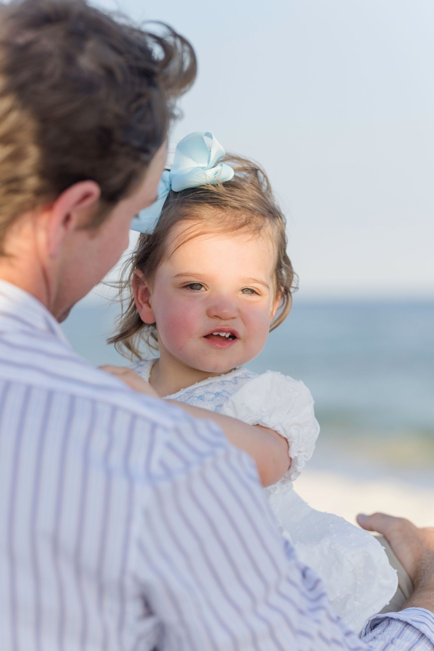 A toddler girl with a blue bow looks at the camera while being held by an adult near the ocean.