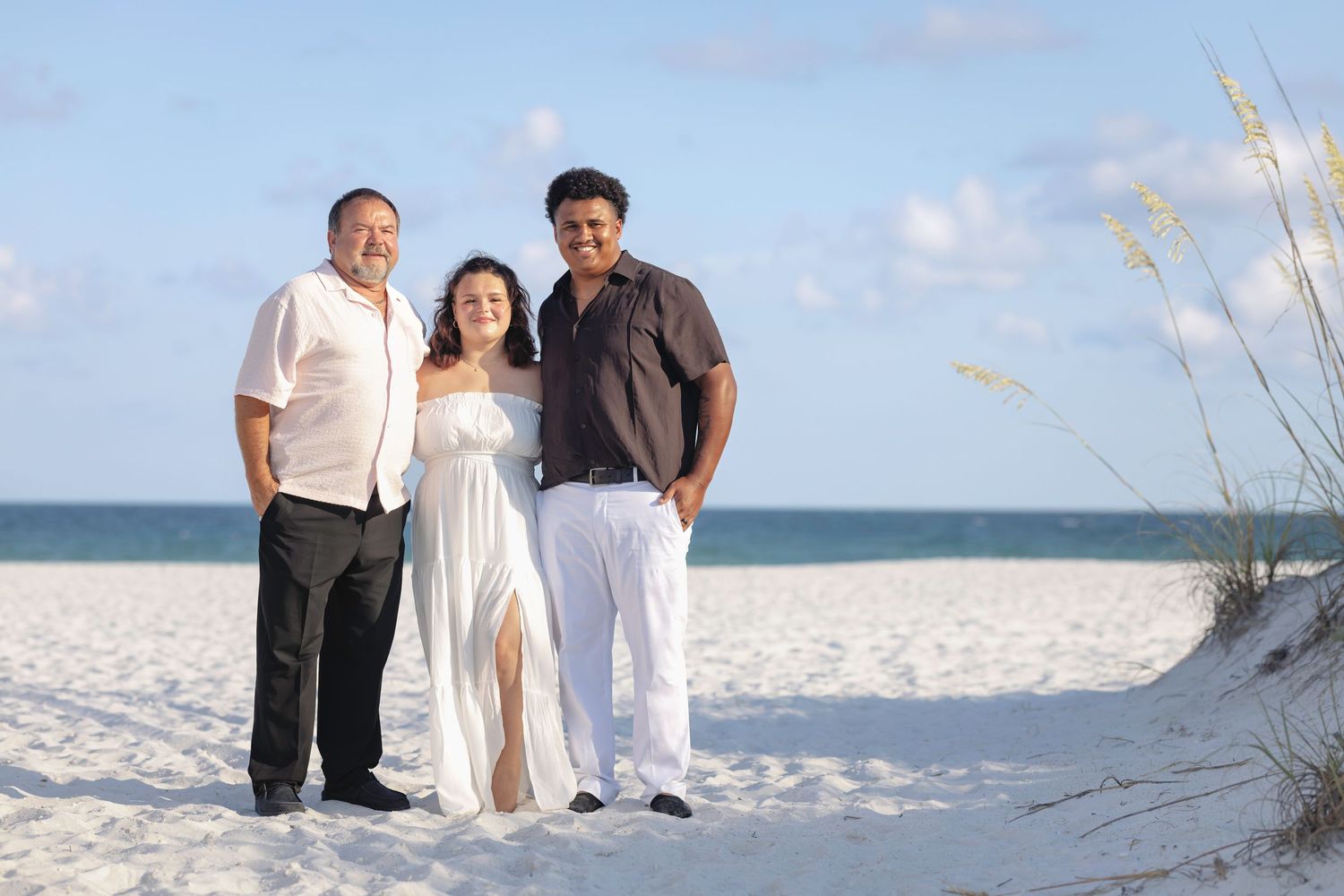 Three people smiling together on a white sandy beach with blue sky and sea oats in the background.