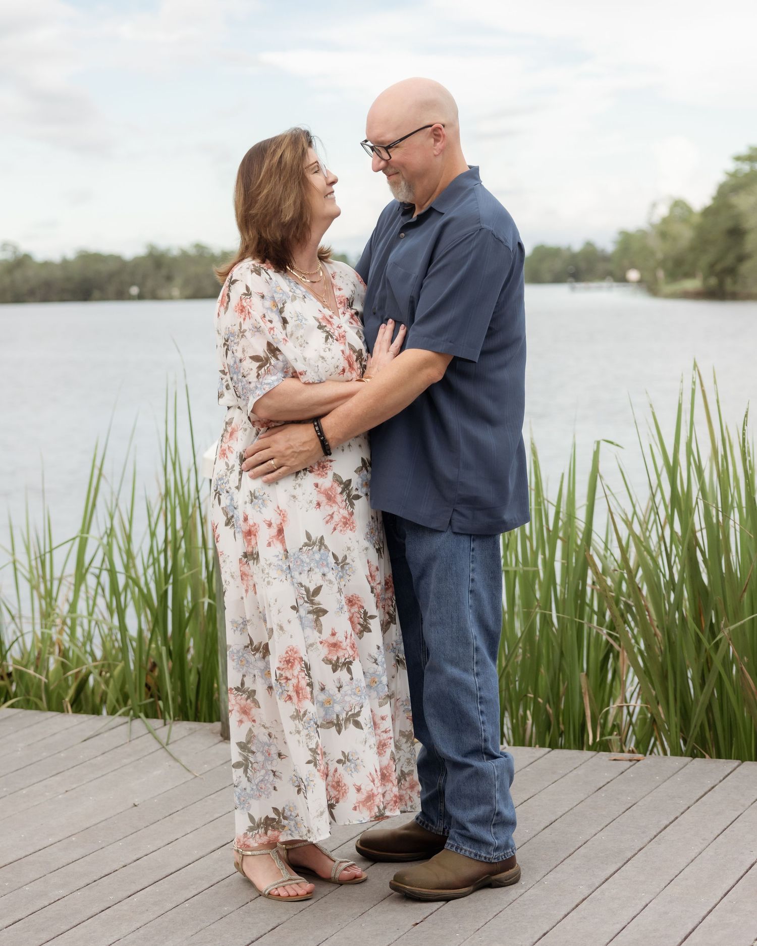 Pregnant couple embracing on a wooden dock by a lake, woman in floral dress, man in blue shirt and jeans.