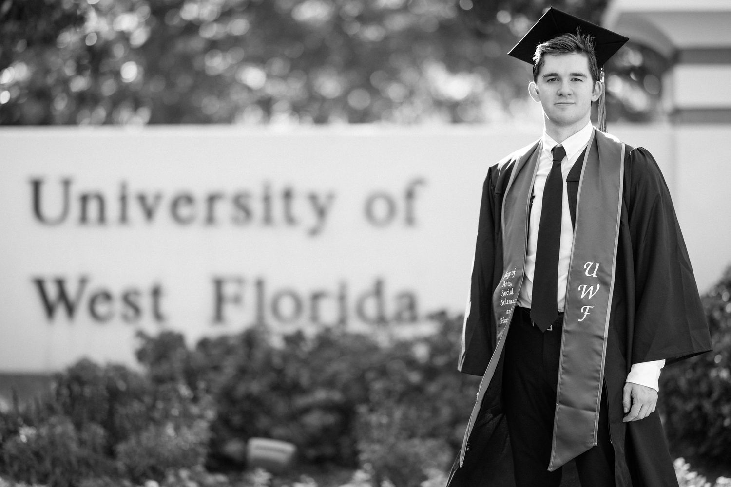 Graduate in cap and gown poses proudly in front of University of West Florida sign in black and white photo.