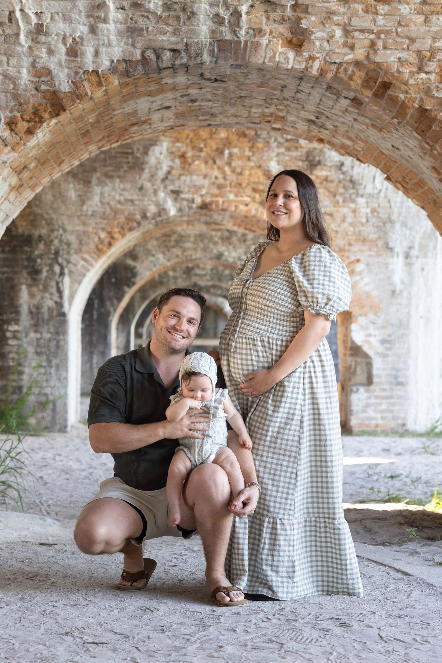 Pregnant woman in gingham dress poses with husband and baby under brick arches at historic fort.