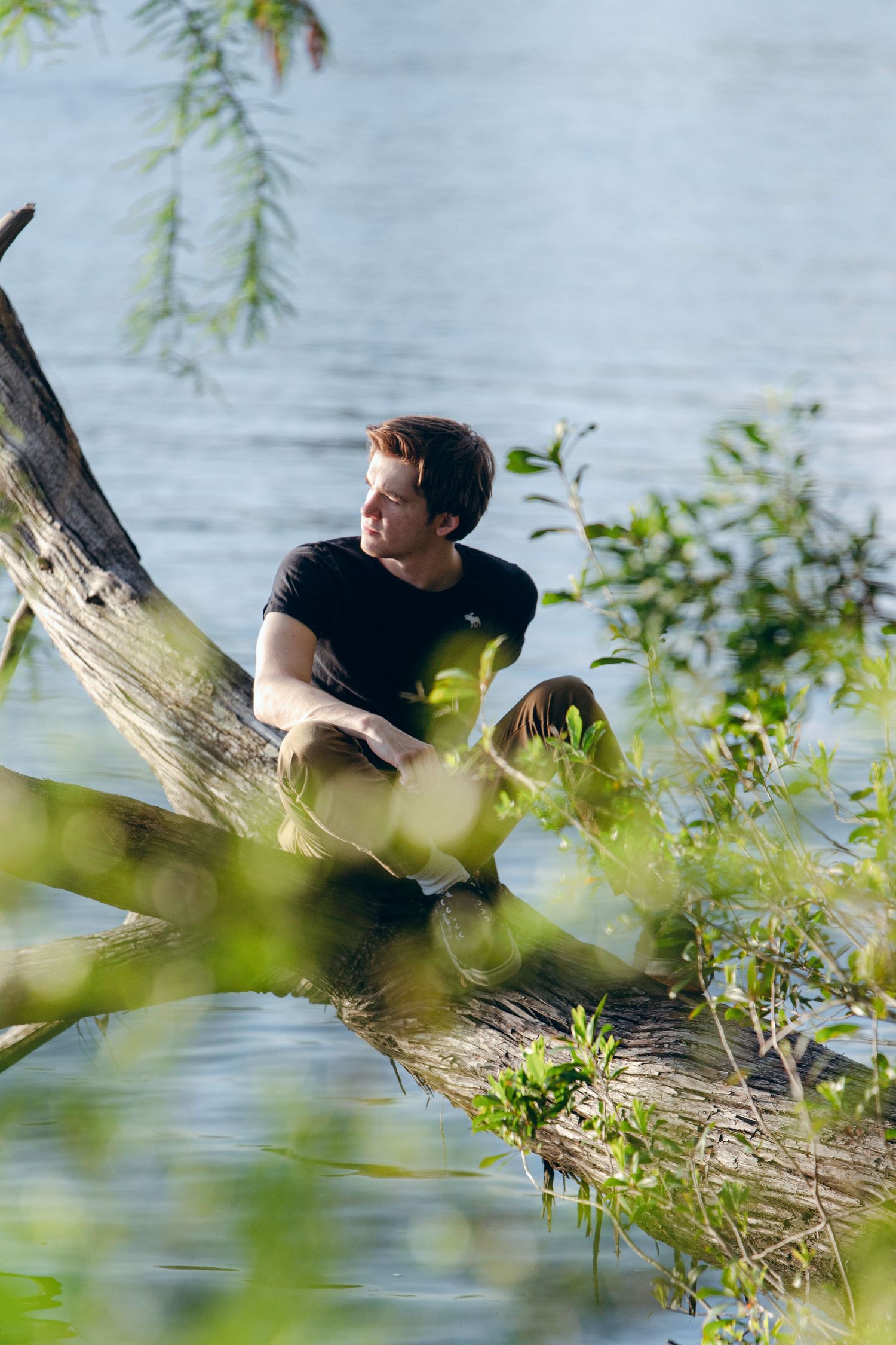 Young man in black t-shirt sitting on a fallen tree trunk by a serene lake, surrounded by lush green foliage.