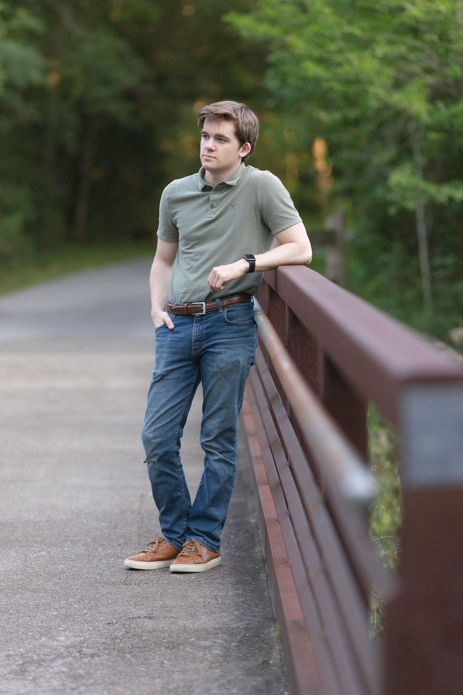 Young man in olive polo and jeans leans on brown bridge railing along a tree-lined path.