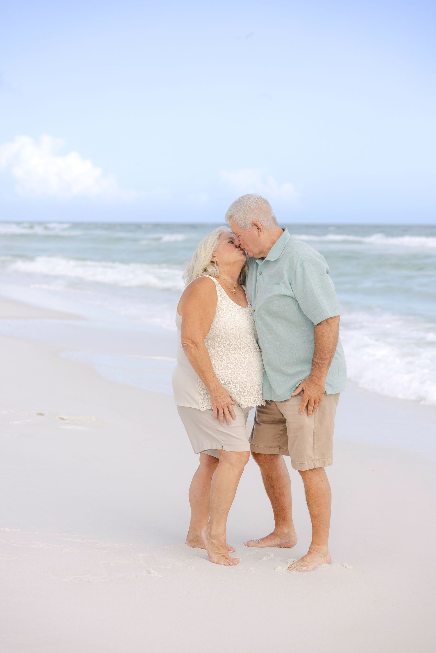 Senior couple in casual beach attire shares intimate moment on sandy shoreline.