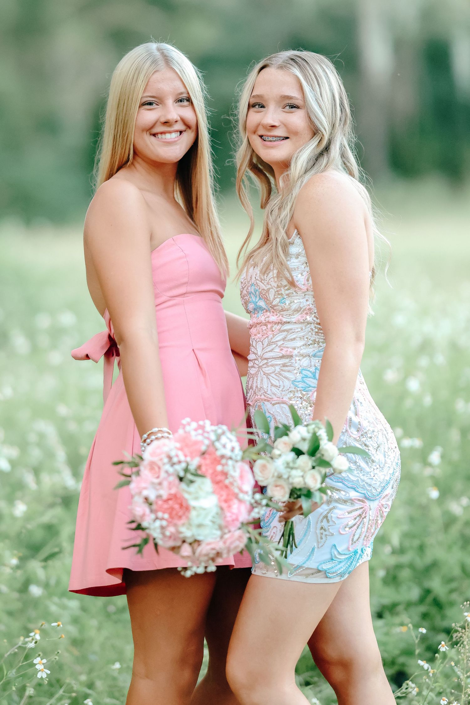 Two smiling blonde women in pink and floral dresses holding bouquets in a sunny flower field.