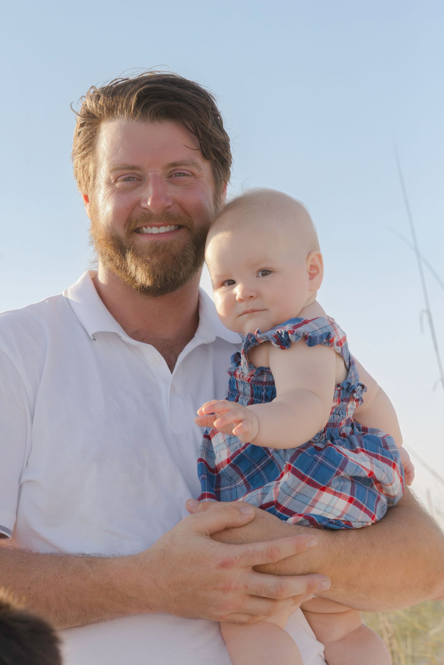 Smiling bearded man in white shirt holding a baby girl in plaid dress outdoors at the beach.