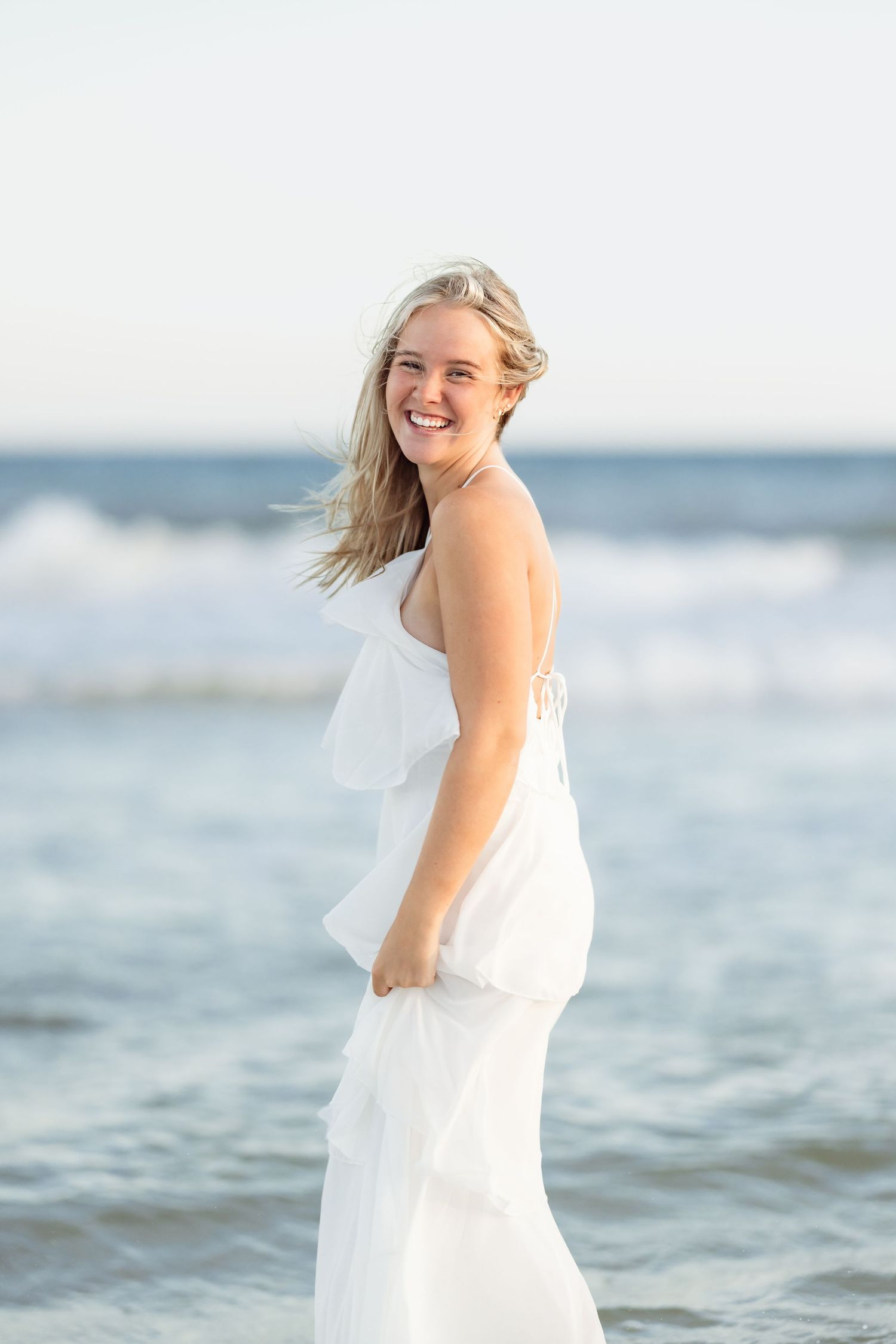 Smiling blonde woman in white dress standing on beach with ocean waves in background.