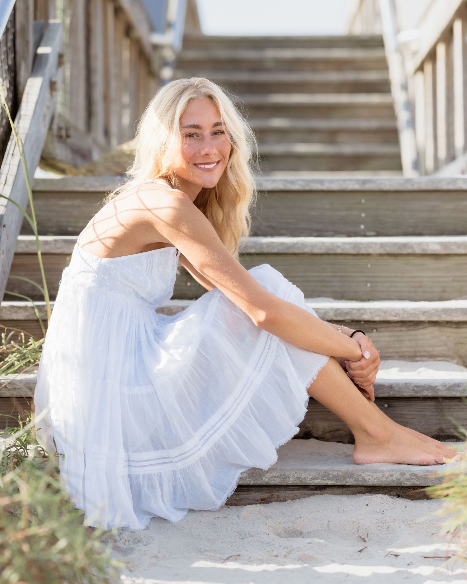 Smiling blonde woman in white sundress sitting barefoot on wooden beach stairs in sunny outdoor setting.