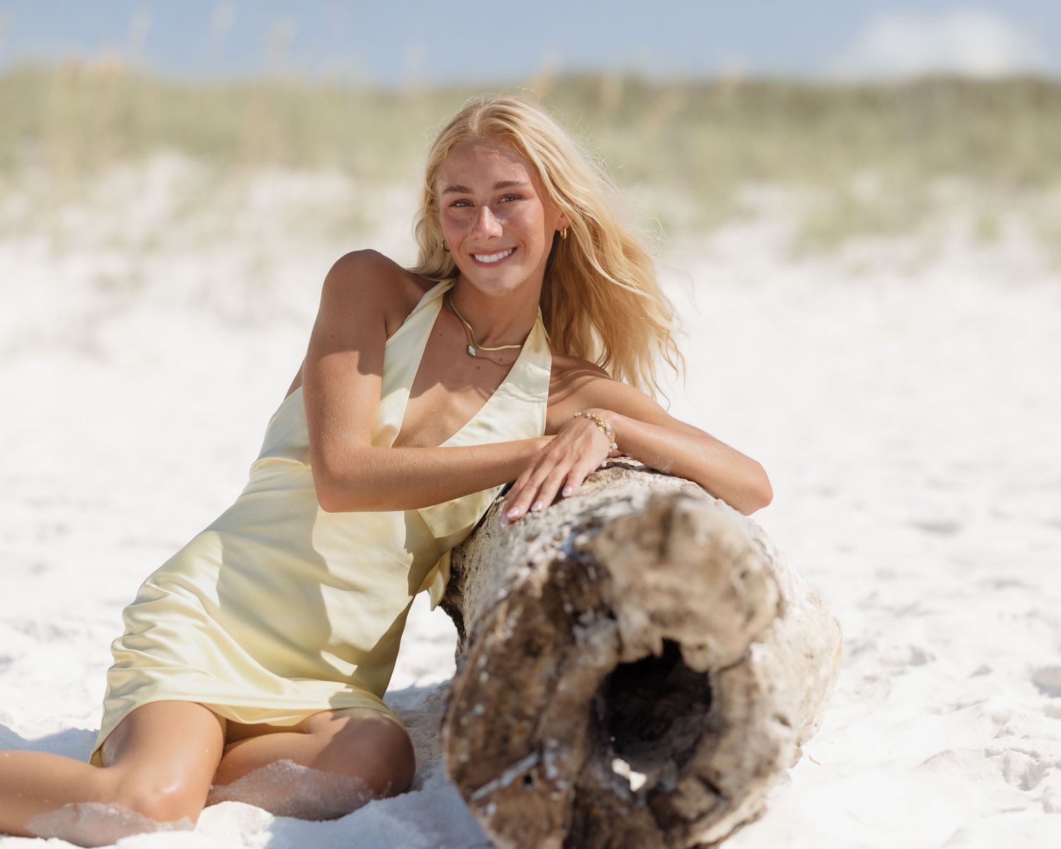 Smiling blonde woman leaning on driftwood on sunny beach.