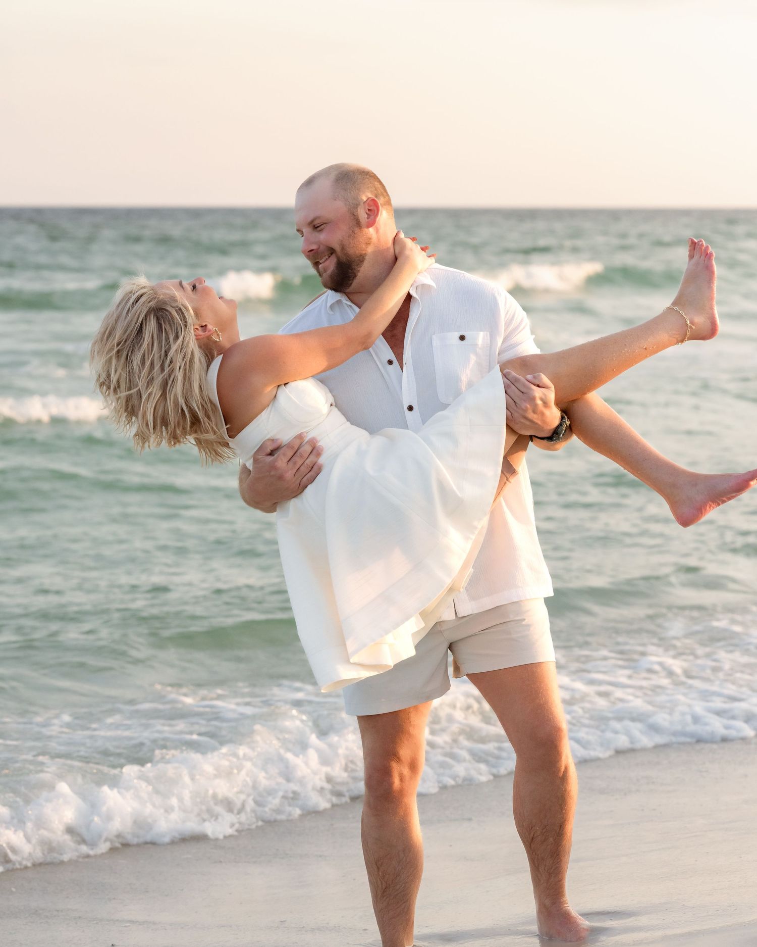 Man in white shirt lifts smiling blonde woman in his arms at the ocean's edge during sunset.