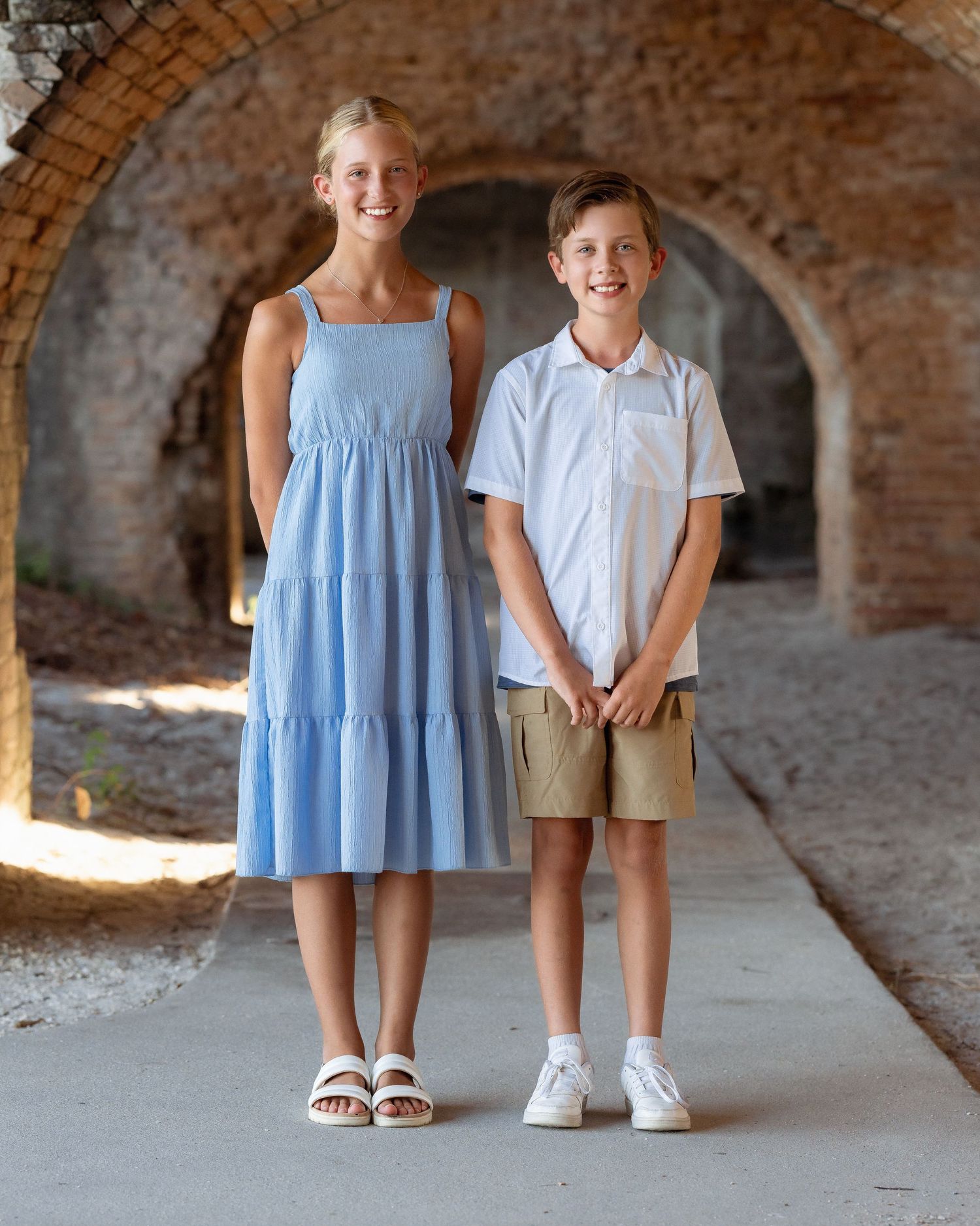 Two smiling children pose in a stone archway, girl in blue tiered dress and boy in white shirt with khaki shorts.