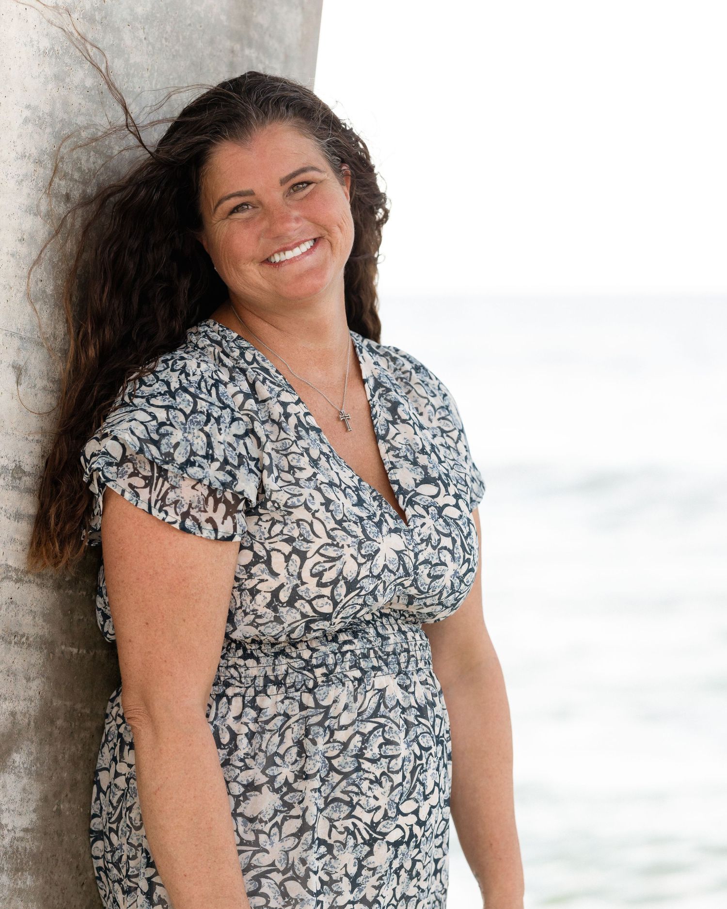 Woman in floral dress smiling at beach leaning against palm tree with ocean in background.