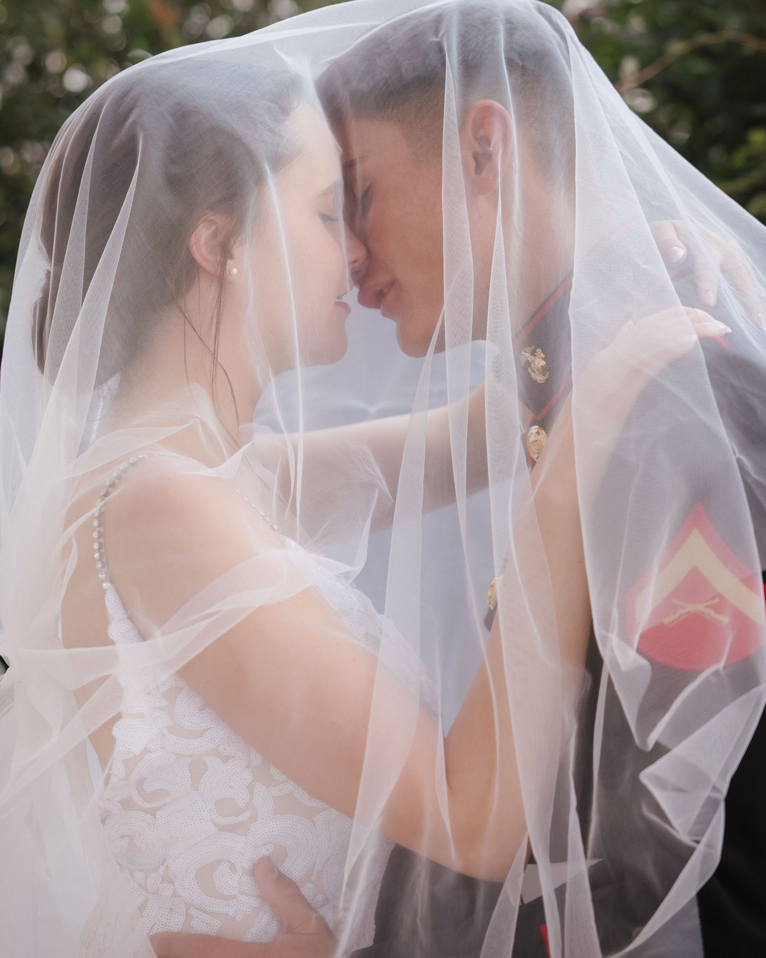 Bride and groom share an intimate moment under a flowing white wedding veil, nearly kissing in a romantic outdoor setting.