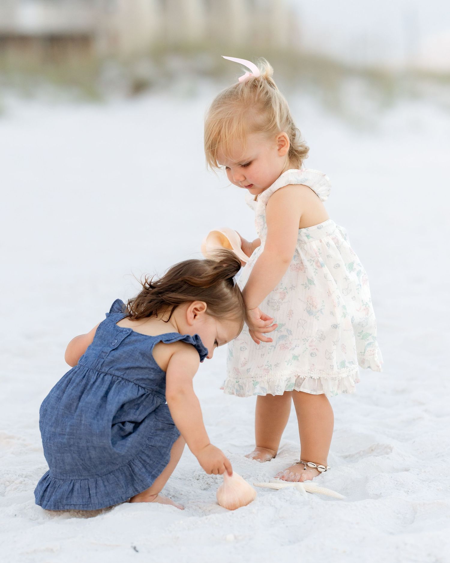 Two toddler girls in dresses playing with a seashell on a white sandy beach.