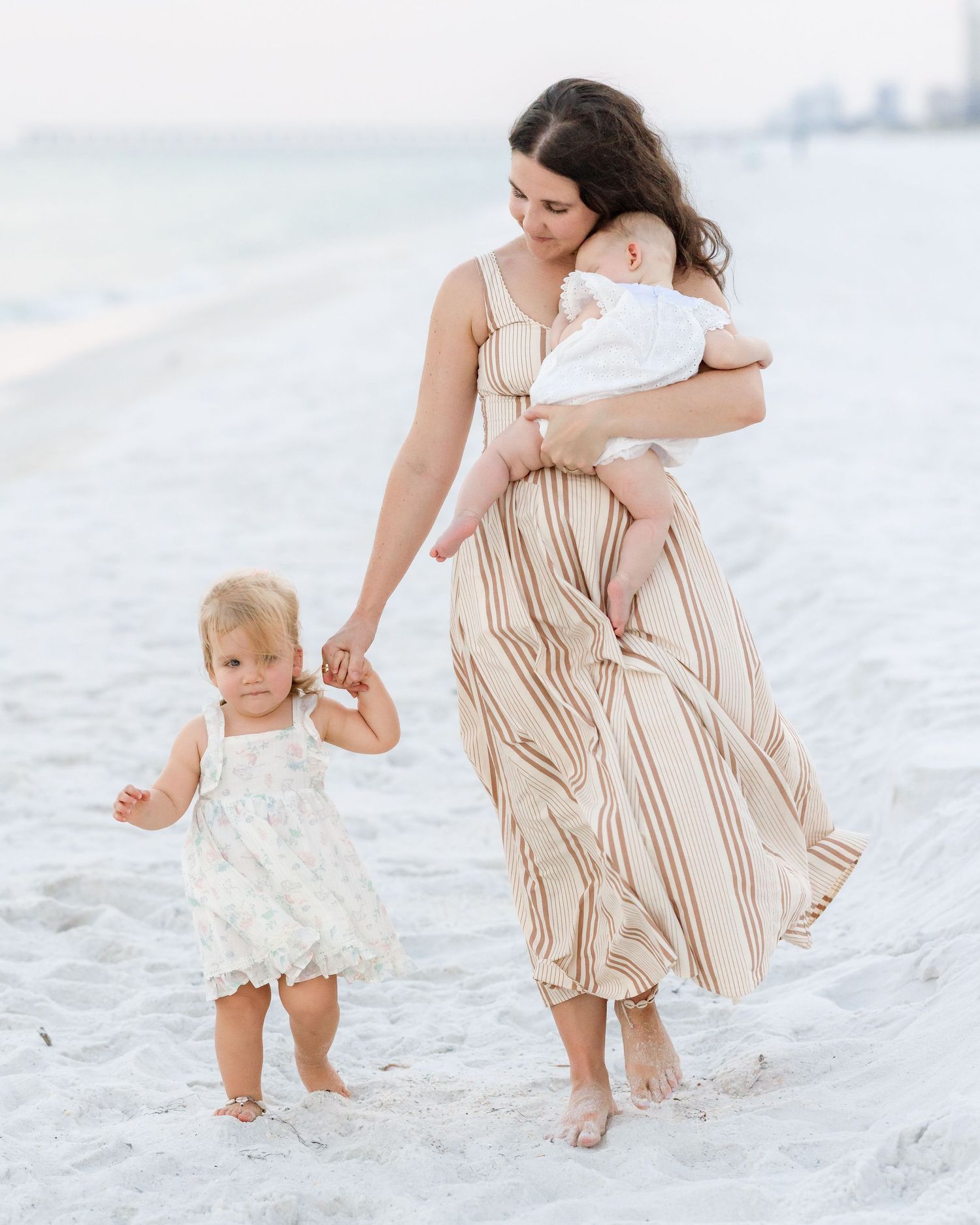 Mother in striped maxi dress holds baby while toddler girl walks beside her on sandy beach.