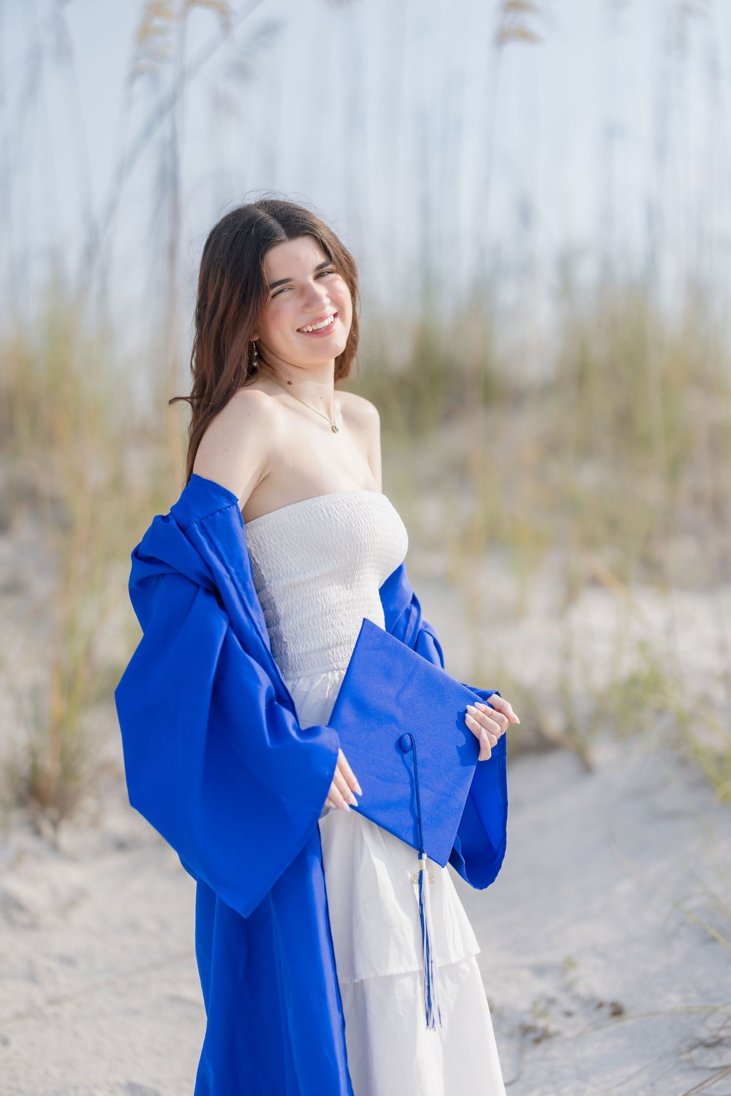 Smiling graduate in blue cap and gown over white dress posing on sandy beach dunes.