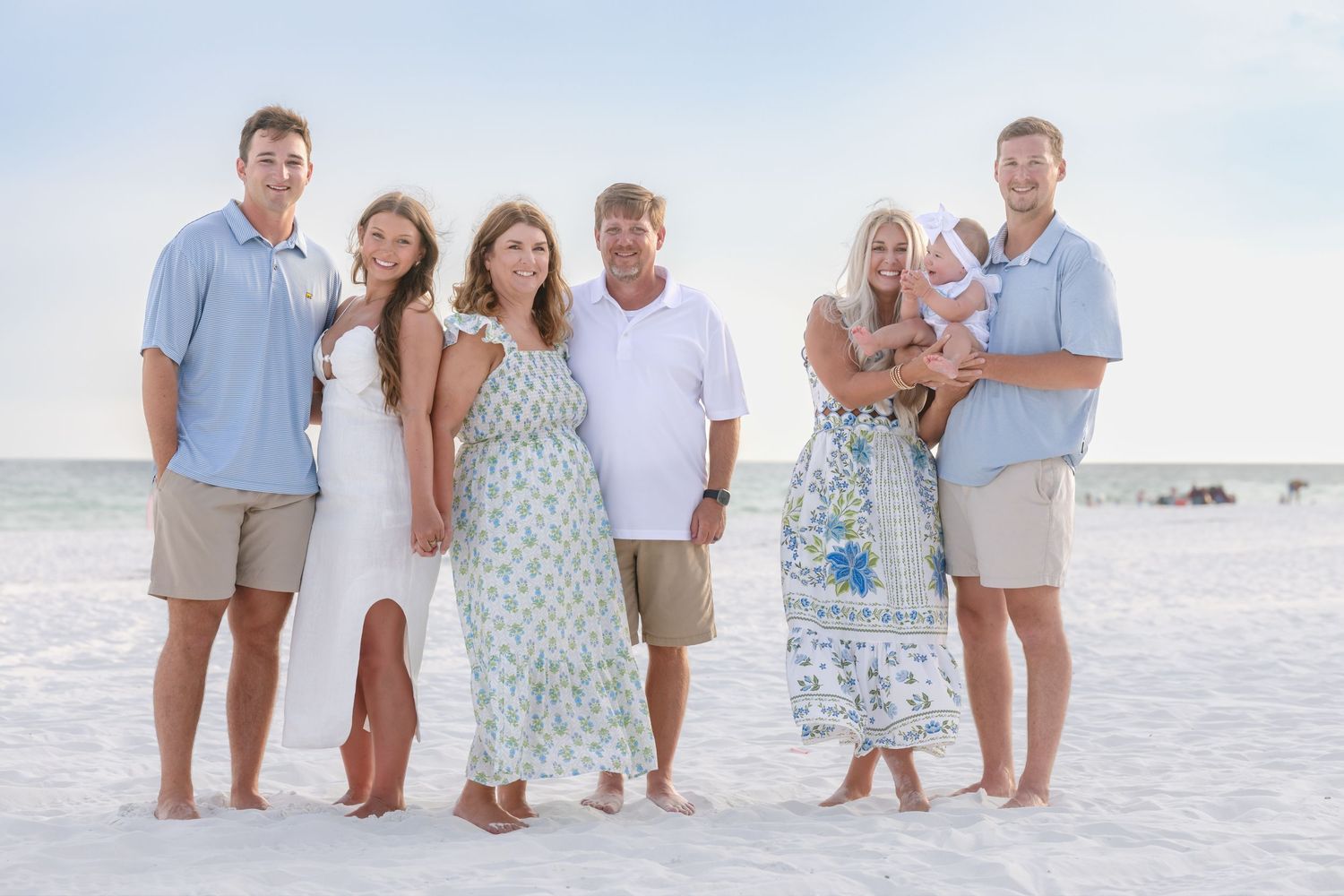 Family of seven posing together on a sandy beach, dressed in coordinating blue and white outfits, holding a baby.