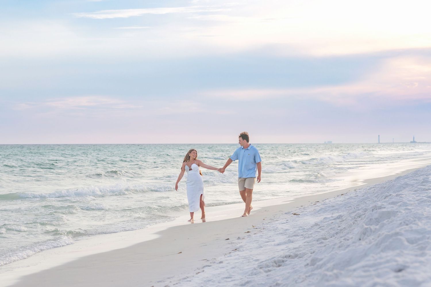 Couple holding hands walking along a white sandy beach at sunset with turquoise waves.