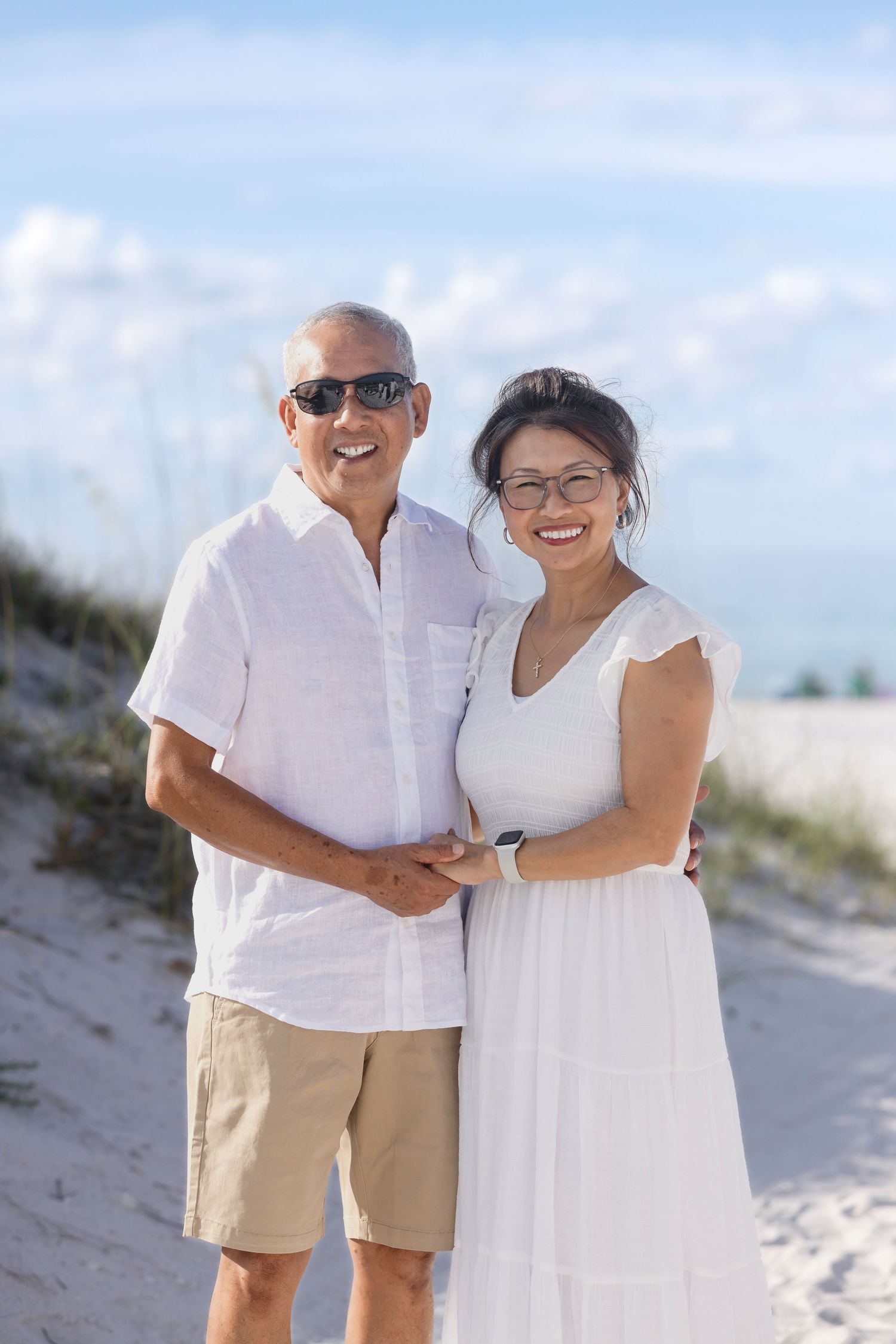 Happy senior couple in white outfits smiling on a sunny beach with sand dunes and blue sky.