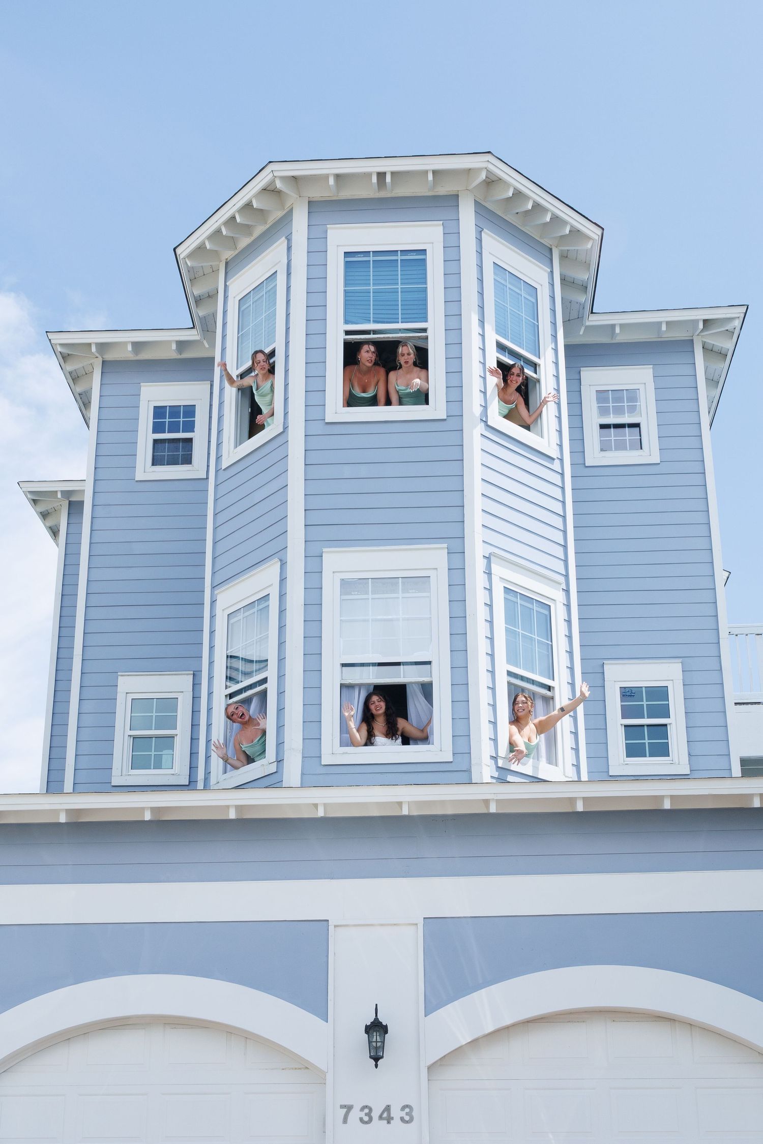 Wedding party members waving from windows of blue three-story beach house numbered 7343.
