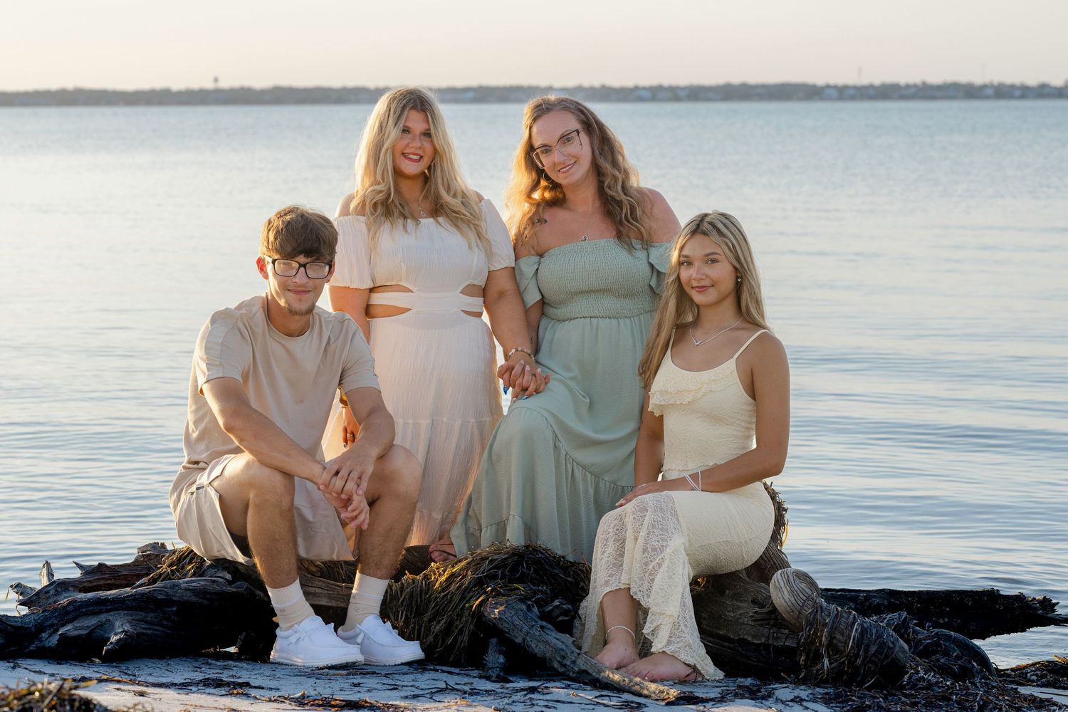 A group of friends in light-colored clothing sit together on rocks near the ocean during golden hour.