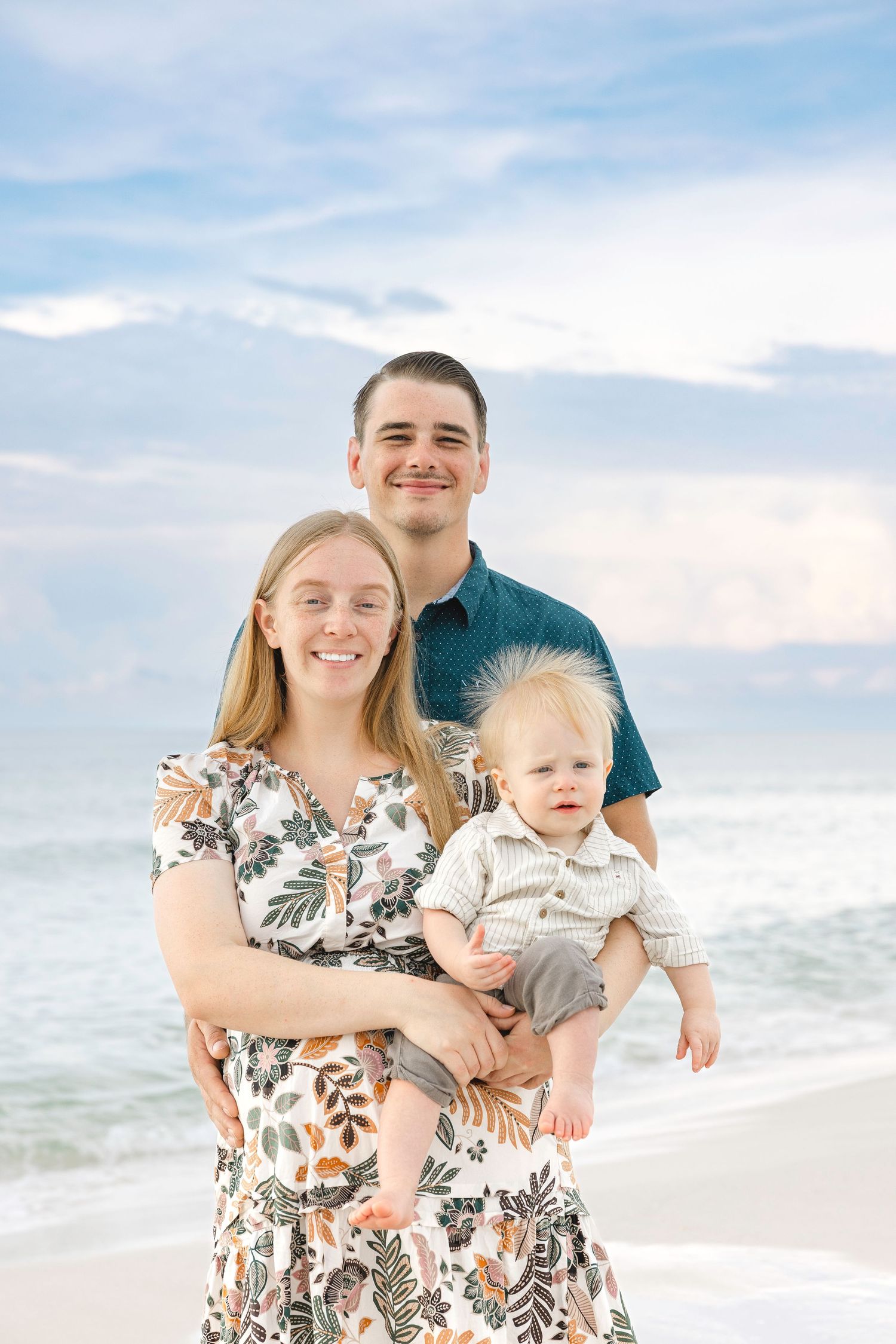 A family of three enjoys a beach photoshoot in coordinated outfits with ocean waves behind them.