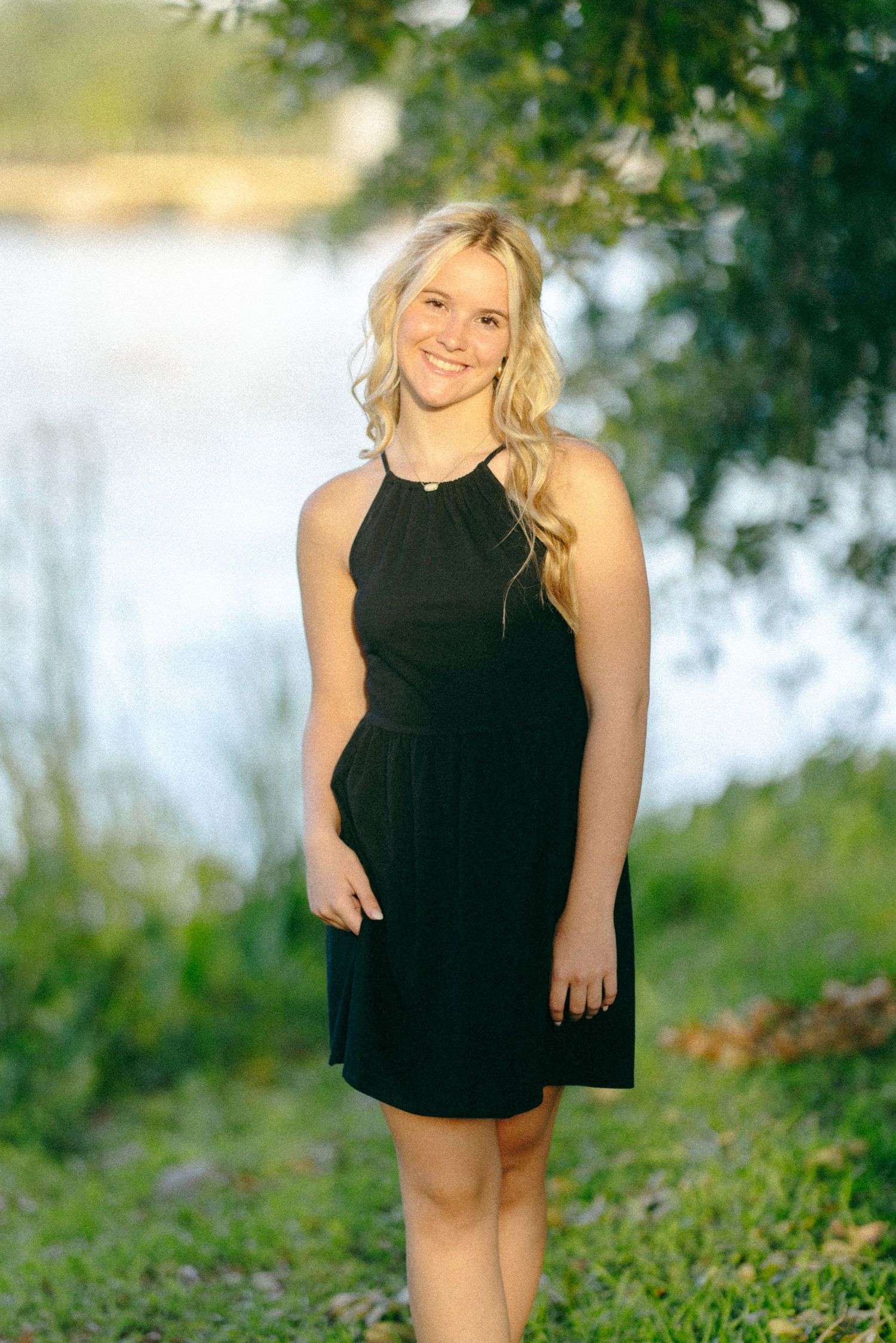 A person wearing a black halter dress poses outdoors against a blurred nature background with water and trees.