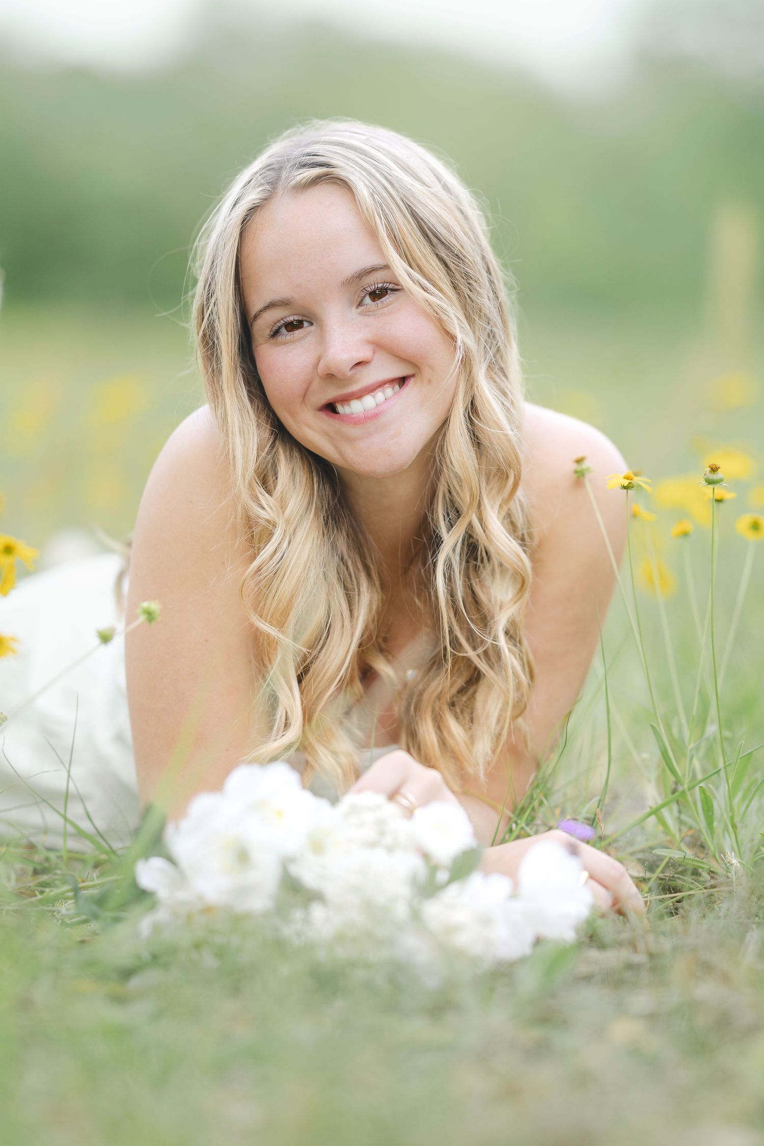 A person lying in a field of white and yellow wildflowers on a sunny summer day, smiling warmly at the camera.